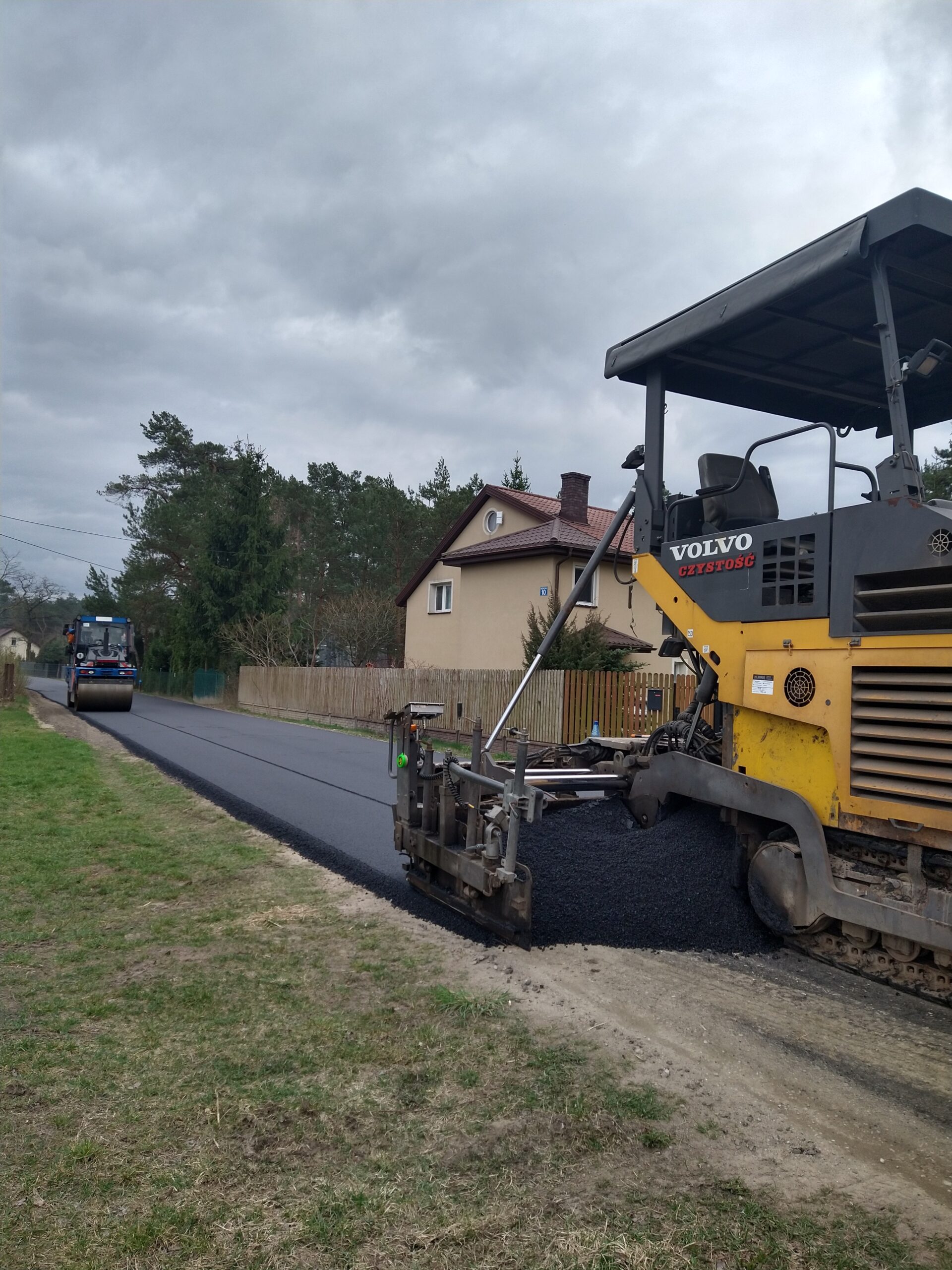 Na obrazku maszyny do układania nawierzchni marki Volvo, która wykłada świeży asfalt na drodze w rejonie mieszkalnym. W tle zauważamy domy oraz drzewa.