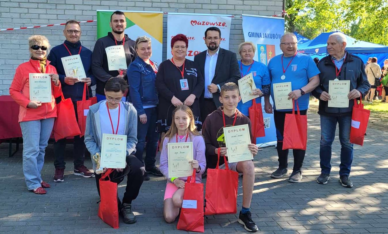 Grupa ludzi stoi na zewnątrz, trzymając certyfikaty i czerwone torby, a następnie mogą mieć medale na szyi. W tle są namioty oraz banery.