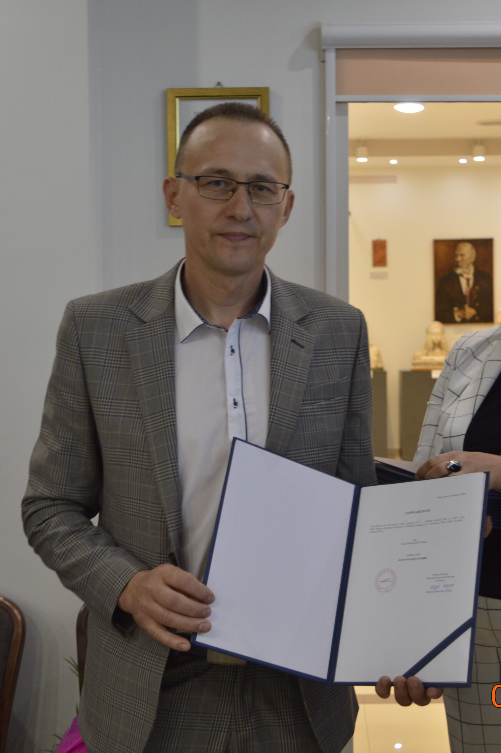 Na obrazku mężczyzn w okularach i w kratkowanym marynarce, który trzyma otwarty segregator. Scena rozgrywająca się w pomieszczeniu.