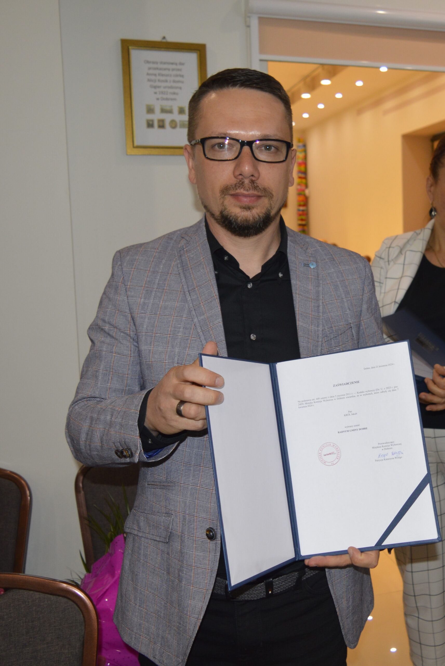 Na zdjęciu jest mężczyzna w okularach i szarym garniturze, który trzyma otwartą teczkę z dokumentami, przechowywaną w pomieszczeniu. Wydaje się być skupionym nad tym, co jest na kartach.