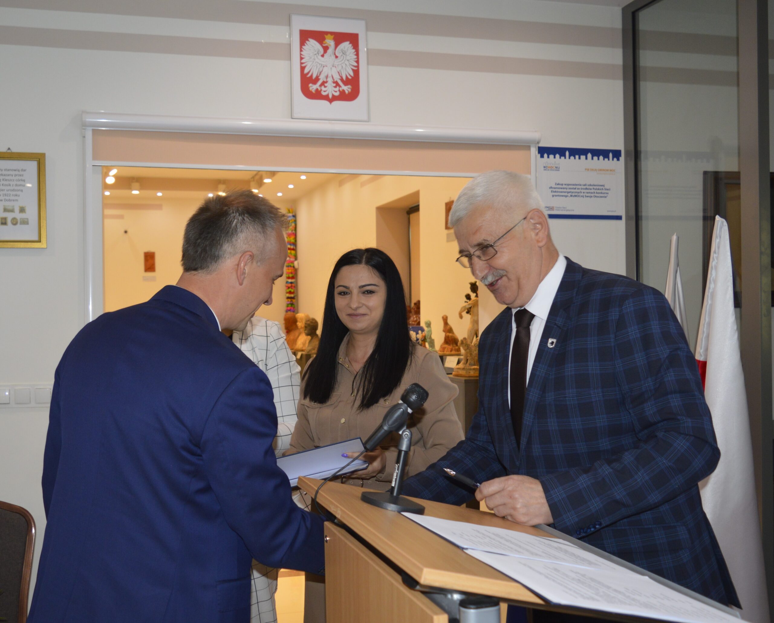 Na obrazku ceremonię z dwóch mężczyzn i jednej kobiety. Jeden z mężczyzn podpisuje dokument na podium z mikrofonem w pomieszczeniu, gdzie jest polska flaga.