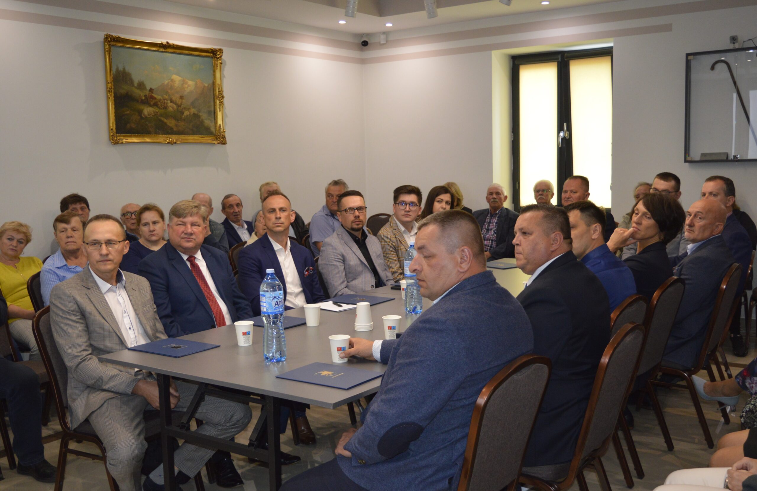 Na zdjęciu grupa dorosłych, która siedzi w sali konferencyjnej i słucha prezentacji. Inne z nich zwróceni są w stronę kamer.