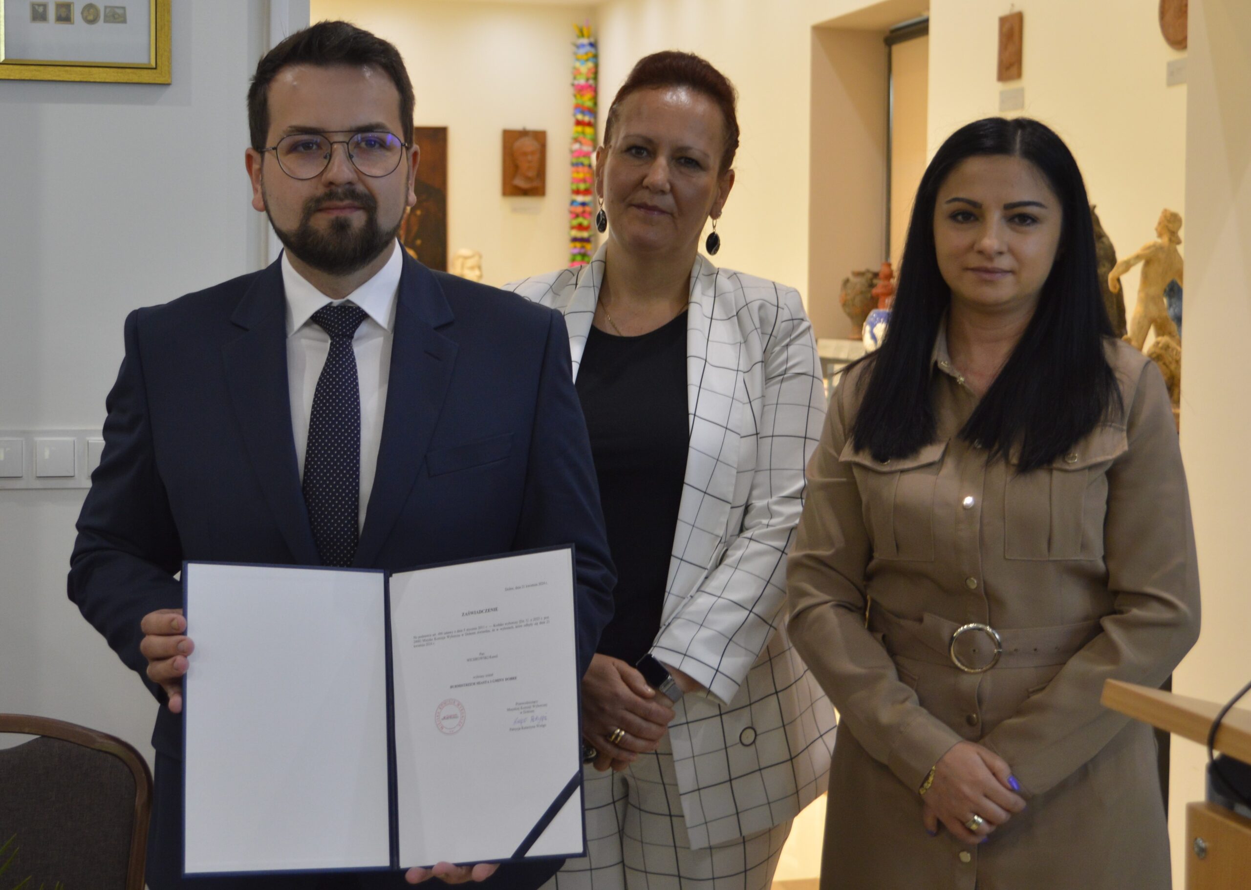Na obrazku widać trzy osoby w pomieszczeniu biurowym. Trzyma otwarty dokument, a z jego obu stron kobiety.