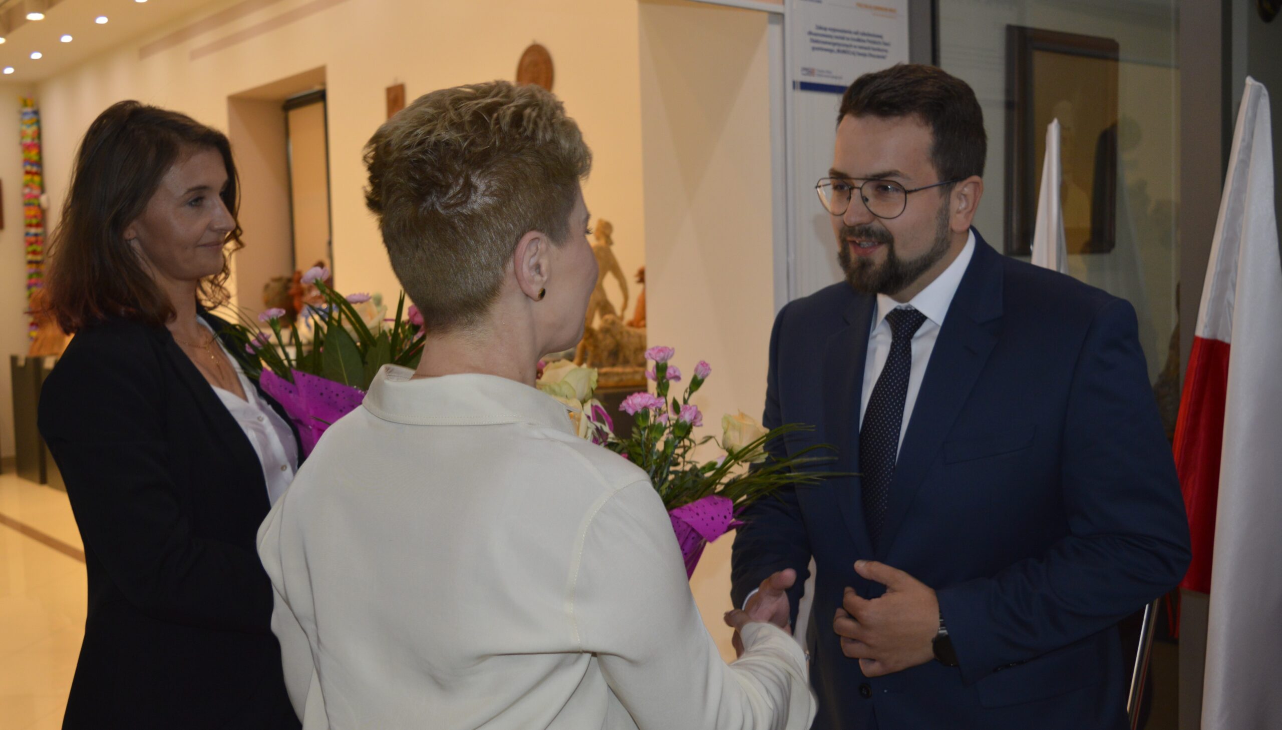 Na obrazku widoczny jest mężczyzna w garniturze, który uścisza dłoń inna osoba ubrana w kremowym stroju, w tle przypominającym galerię. Obok nich kobieta przechowująca bukiet kwiatowy.
