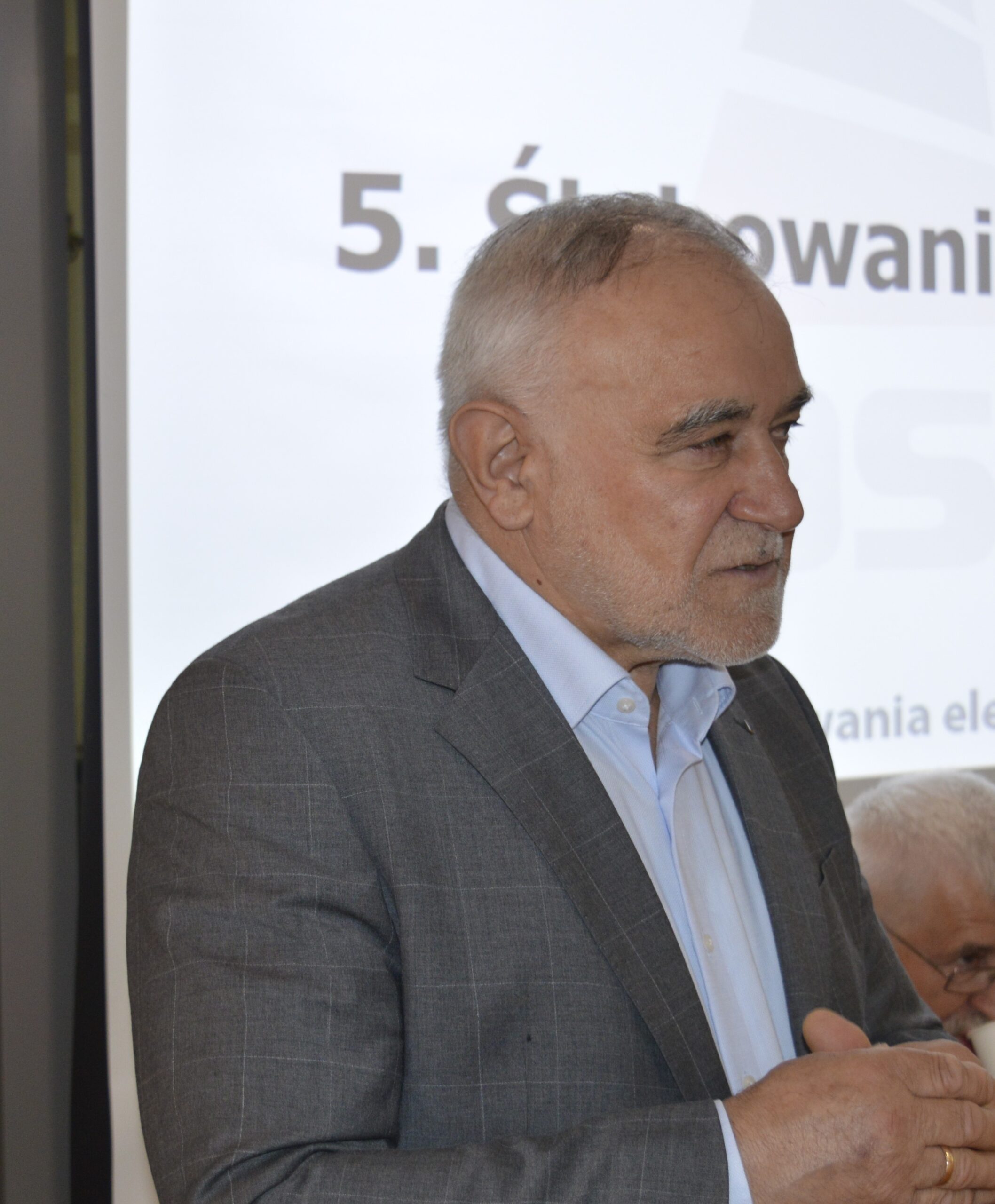Na obrazku dojrzałego mężczyzn o siwych włosach i brodzie, przemawiającego z mównicy. Jest ubrany w szarą koszulę i stoi przed prezentacją.
