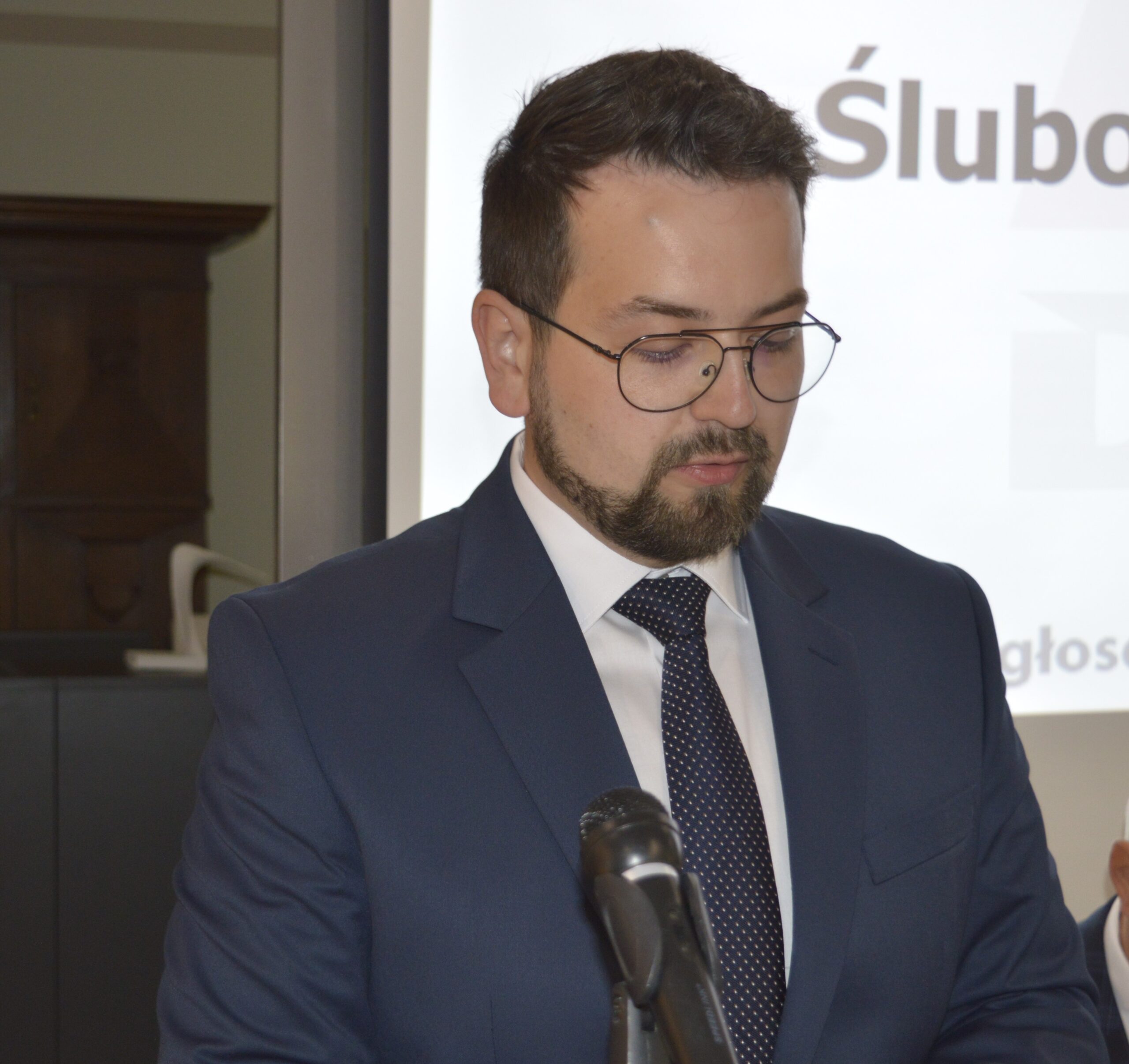 Na obrazku widoczny jest mężczyzna w niebieskim garniturze i okularach, który przemawia do mikrofonu przy mównicy. W tle zauważalny jest ekran prezentacyjny.