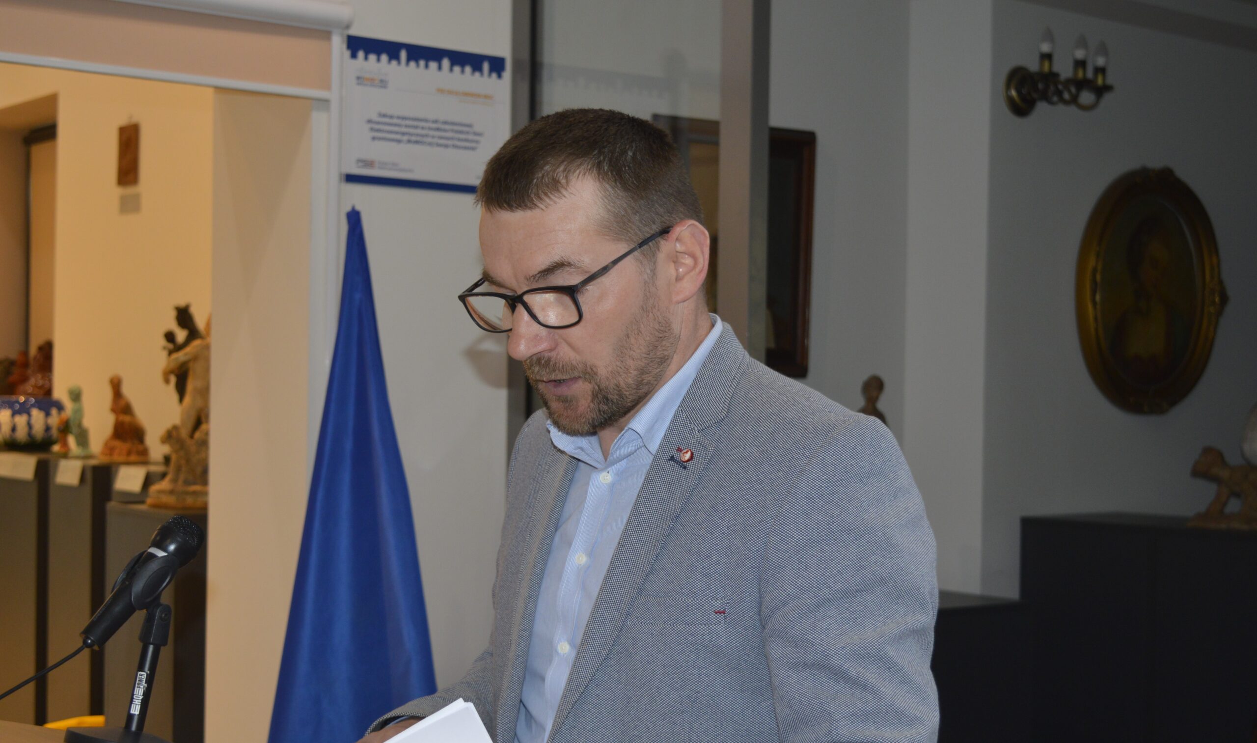 Na widok mężczyzn w okularach i szarych marynarkach, który przemawia przy podium z mikrofonami. W tle znajdują się flagi Unii Europejskiej oraz różne artefakty.