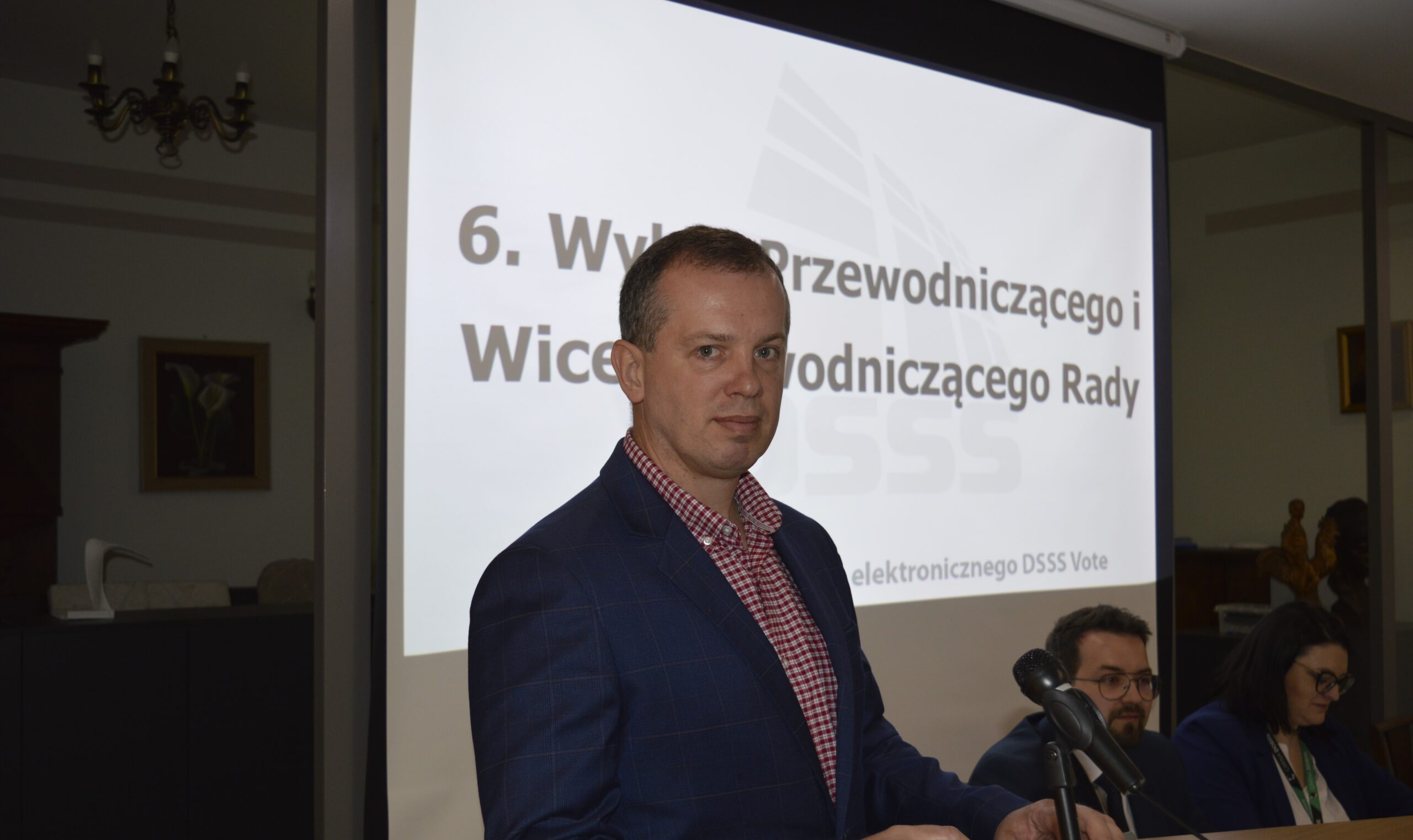 Na obrazku mężczyzn w niebieskim garniturze i czerwonym krawacie, który stoi przed mikrofonem i przemawia do głośnika. W tle jest prezentacja slajdu.