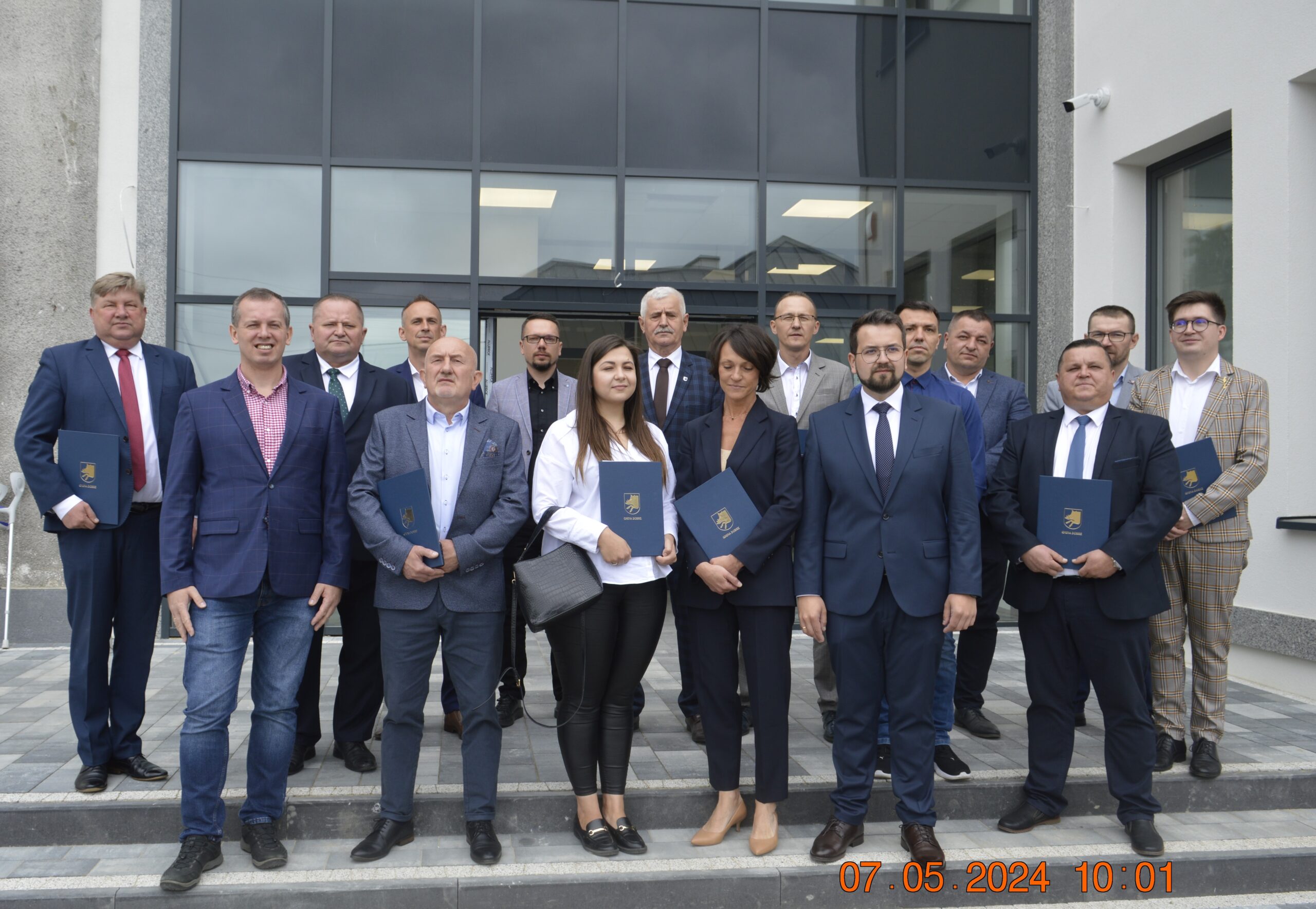 Na zdjęciu posegregowana grupa czternastu konkurencyjnych firm trzymających certyfikaty. Stoją oni na tle budynku i pozują do zdjęć.