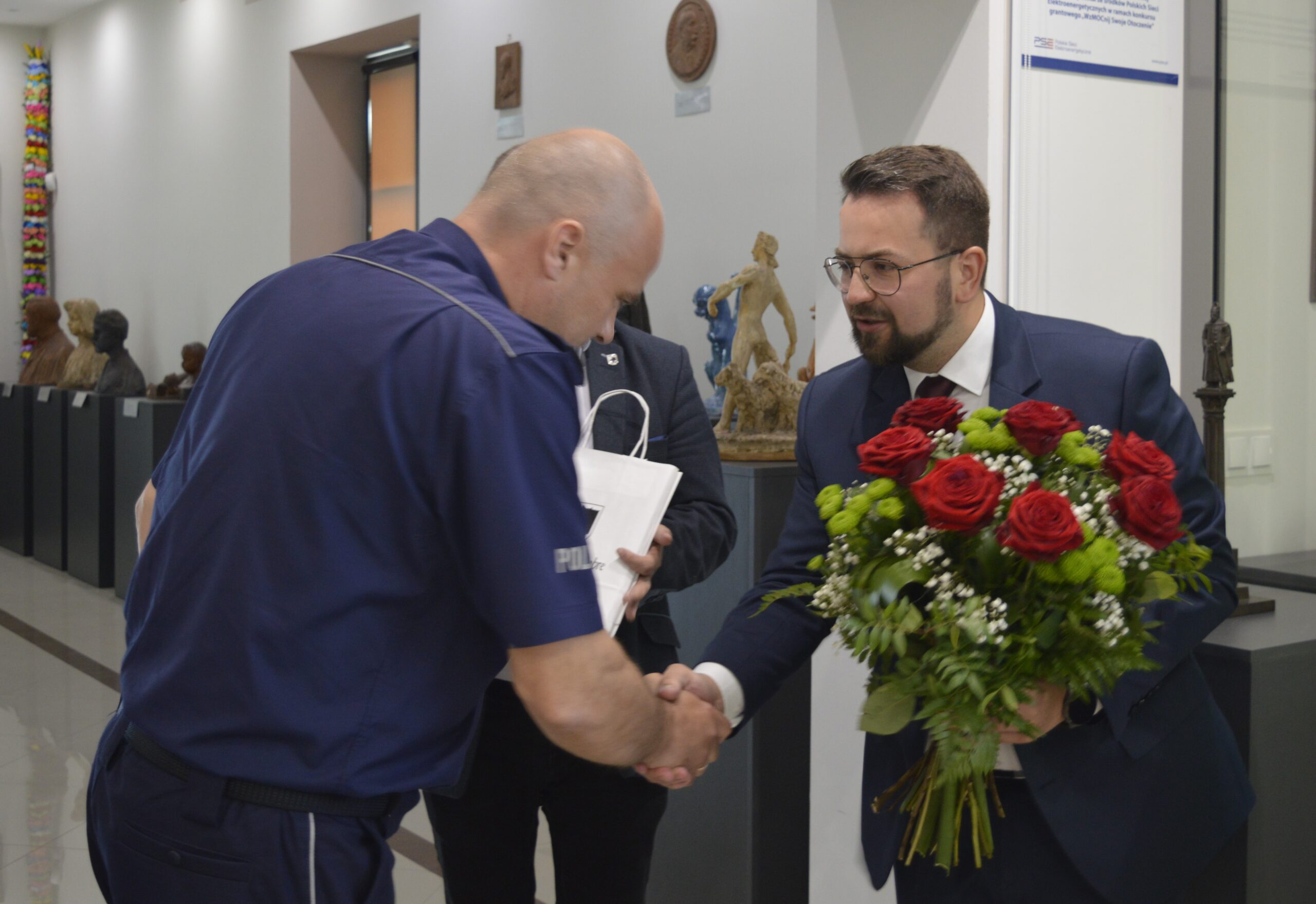Na obrazku mężczyzny w garniturze i okularach ściskanych w dłoni mężczyzn w mundurze, trzymanych przy tym bukietu czerwono-białych kwiatów. Znalezienie się w pomieszczeniu, w którym znajduje się informacja, jest tablicą pamiątkową.