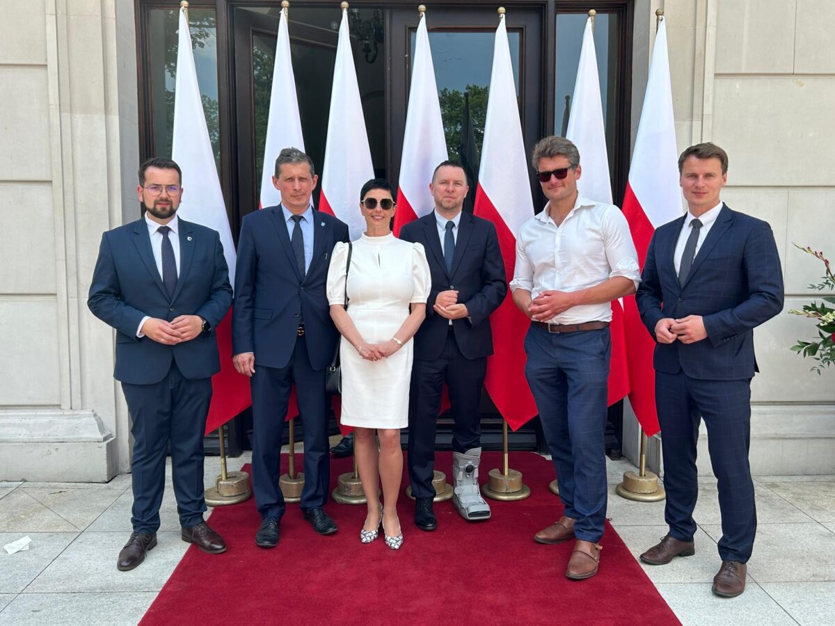 Na sześć osób stoi na czerwonym dywanie przed zestawem białych i czerwonych flag, ubranych w stroje formalne. Grupa pozuje do zdjęć przedem z białymi kolumnami.