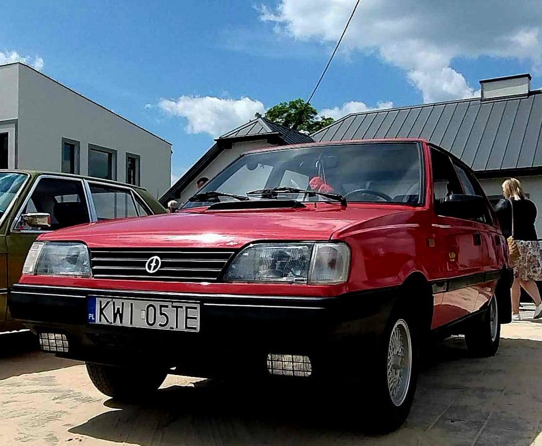 Na zewnątrz znajduje się czerwony samochód z tablicą rejestracyjną „KWI 05TE”, wprowadzony na zewnątrz w słoneczny dzień. W tle widoczne są kilka budynków oraz inny pojazd.