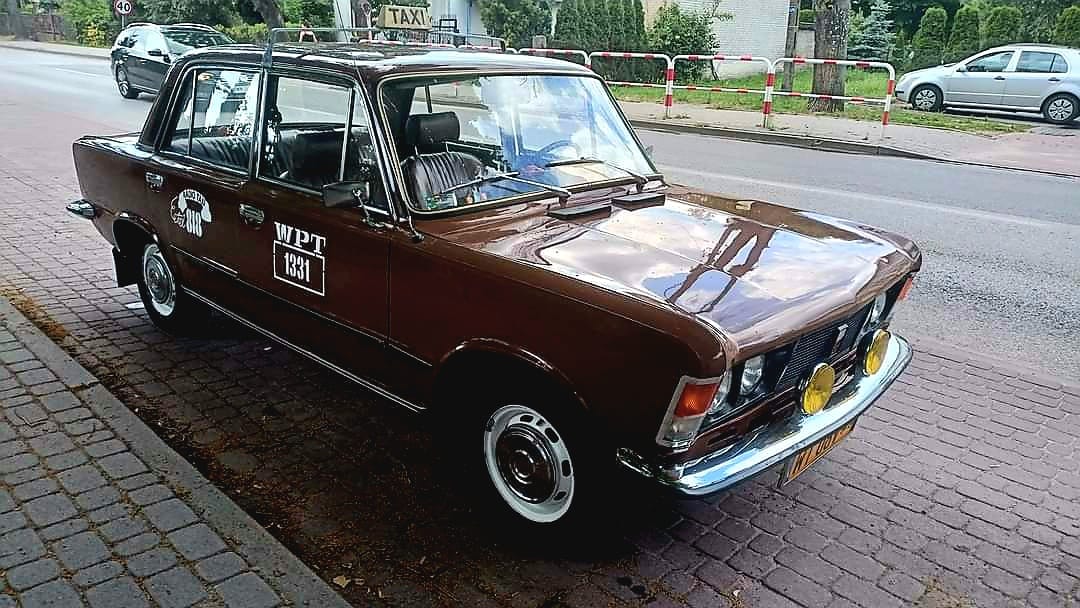 Na ulicy znajduje się zabytkowy brązowy samochód z naklejką „WPT 1313”, umieszczony na ulicy, wyposażony w żółte światła przeciwmgielne oraz znak TAXI na dachu. W tle widać srebrny samochód oraz chodnik drzewami.