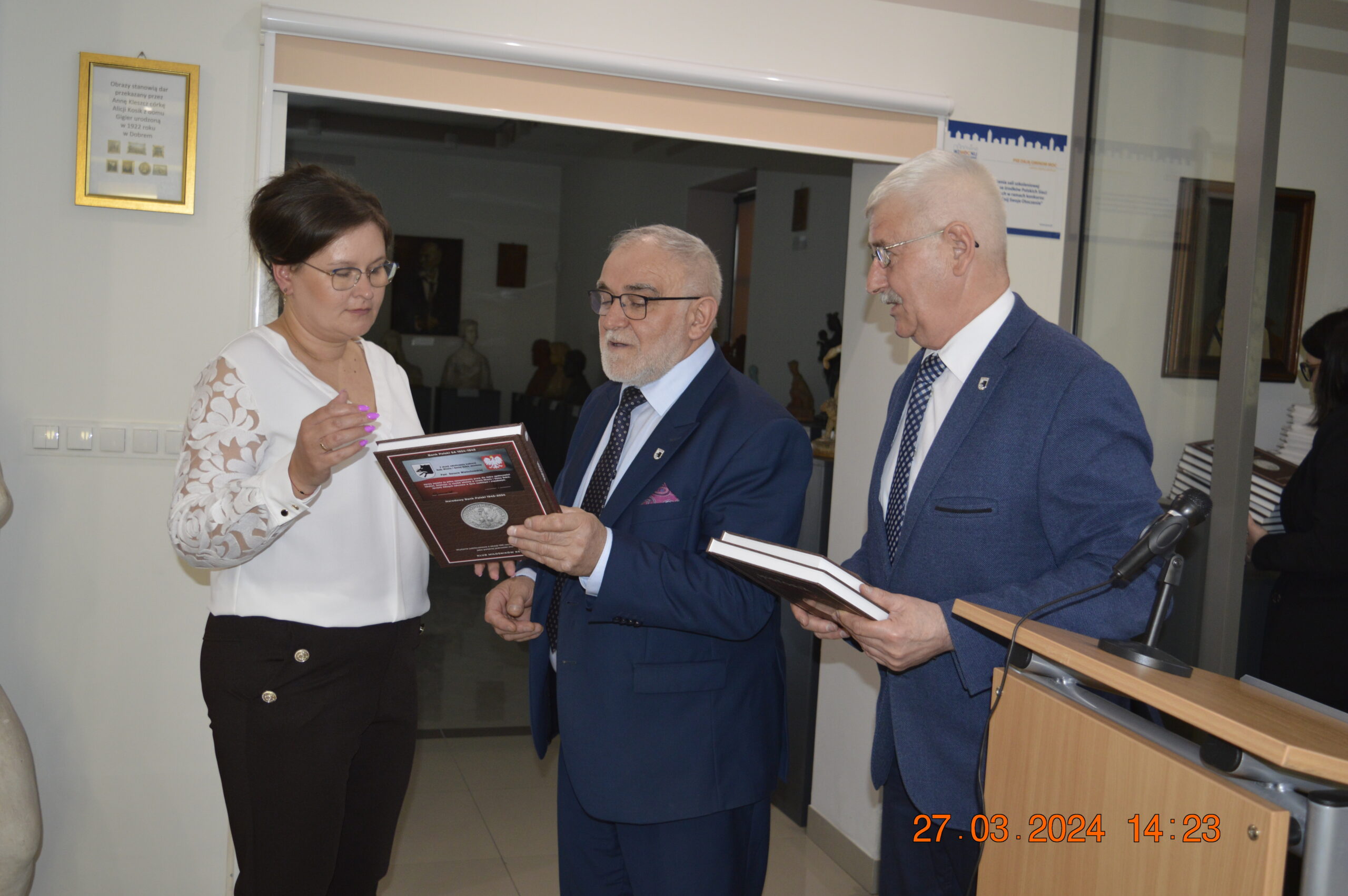 Na obrazku znajdują się trzy osoby ubrane w formalnych ubraniach, które są częścią wnętrza budynku. Kobieta po lewej stronie trzyma plakietkę, a dwóch mężczyzn obok niej wygląda na to, że jest ona bezpośrednio trzymana i jednocześnie trzyma ją. Na zdjęciu widnieje również stempel czasowy z inspektorem „27.03.2024 14:23”.