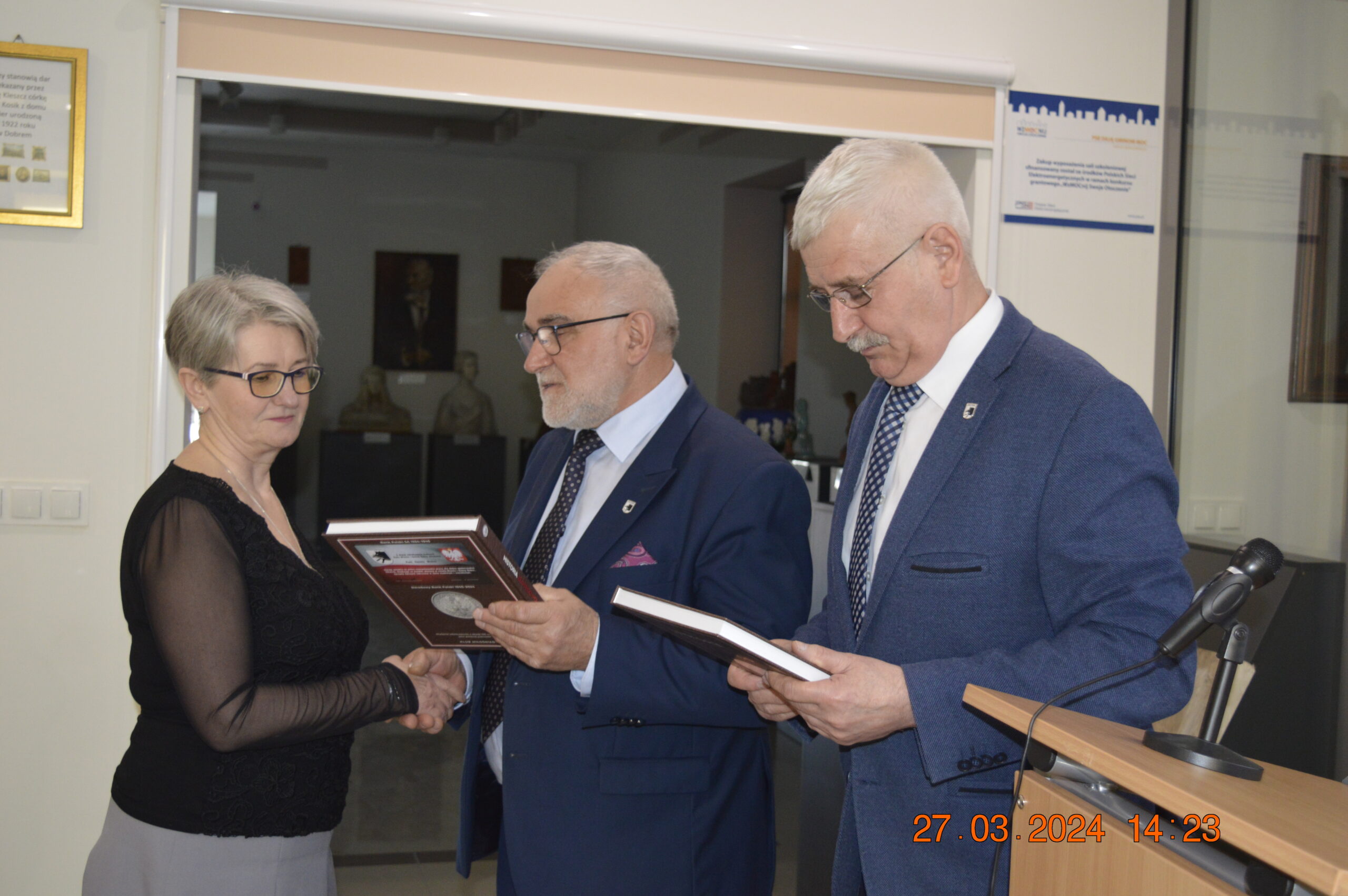 Na zdjęciu trójka osób w eleganckich ubraniach stojących wewnątrz pomieszczenia; kobieta trzymana w ręce, podczas gdy dwóch mężczyzn przegląda dokumenty lub inne. W osobnym dolnym rogu jest data „27.03.2024”.
