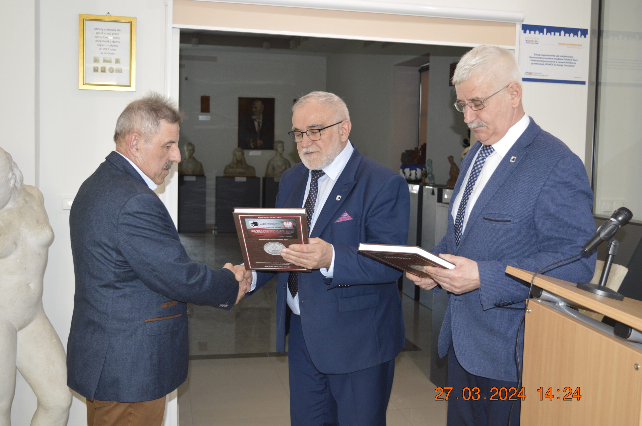 Na obrazku trzech mężczyzn w garniturach, z których dwaj trzymają plakietki. Jeden z nich przekazuje plakietkę drugiemu, ściskając mu rękę, a w tle pojawia się podium i statuetka. Dane wyświetlona na zdjęciu do 27 marca 2024 roku, godzina 14:24.