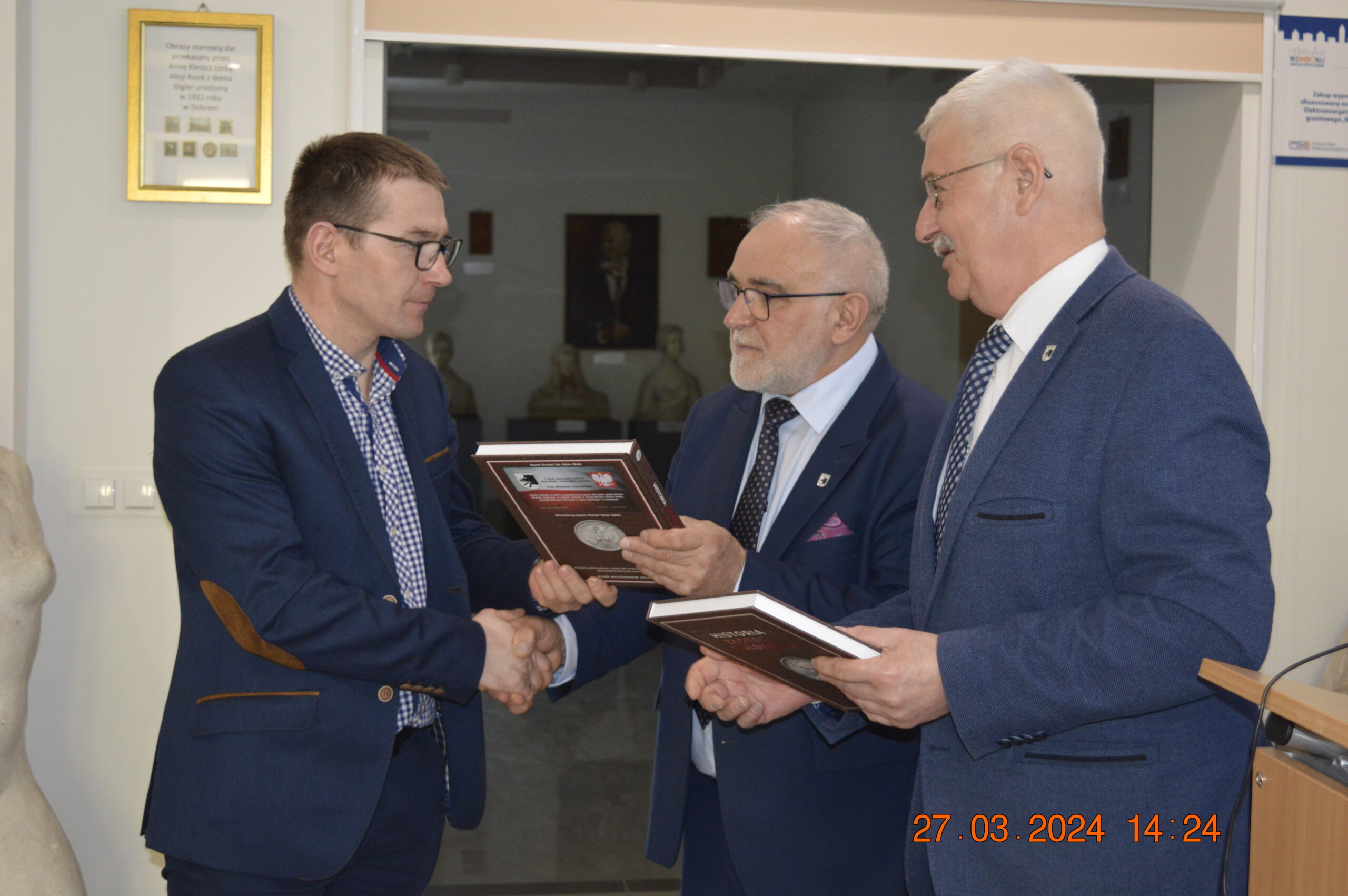 Na obrazku trzech mężczyzn w garniturach wymieniających uściski dłoni oraz trzyma plakiety, co ważne formalną lub wręczenie nagród. W odpowiednim dolnym rogu widnieje dane „27.03.2024” i godzina „14:24”, wydarzenie moment tego wydarzenia.