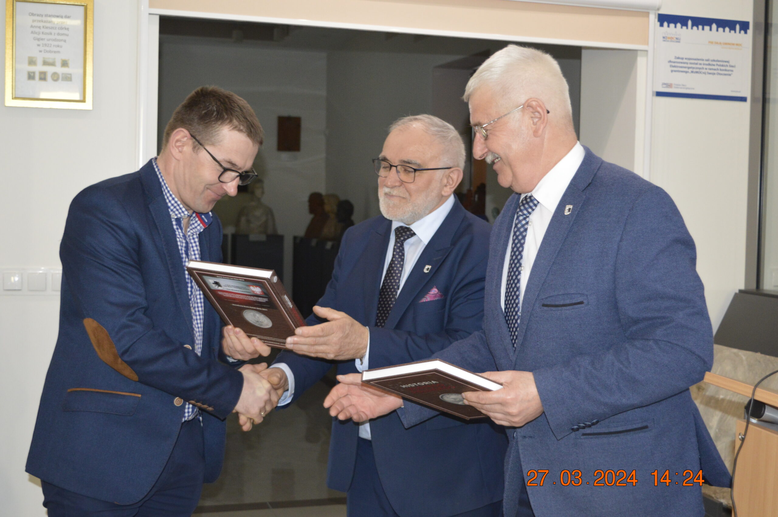Na obrazku trzech mężczyzn w garniturach wymienia się dokumentami i daje sobie ręce w pomieszczeniu. W tle widać zegar, który zawiera datę i godzinę: 27 marca 2024 roku o 14:24.