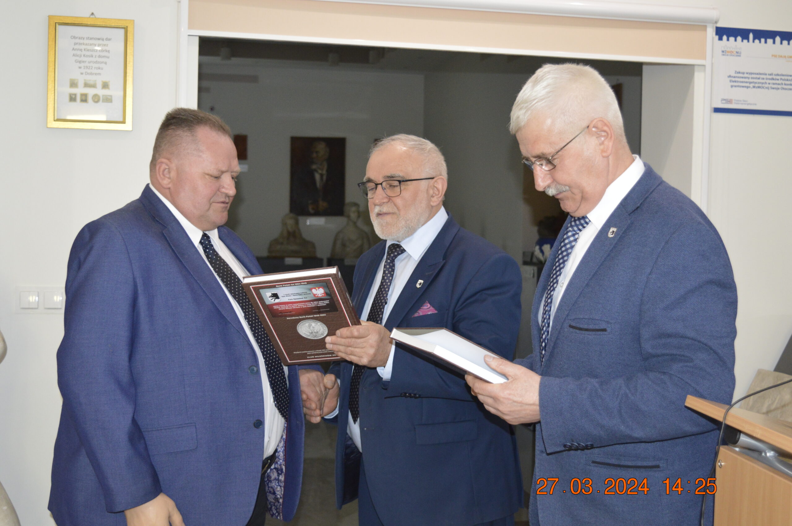 Na obrazku widać trzech mężczyzn w niebieskich garniturach, którzy przeglądają dokumenty w pomieszczeniu. Jeden z nich trzyma oprawiony przedmiot, podczas gdy pozostają dwaj analizują; na zdjęciu widnieje data i godzina: 27.03.2024 o 14:25.