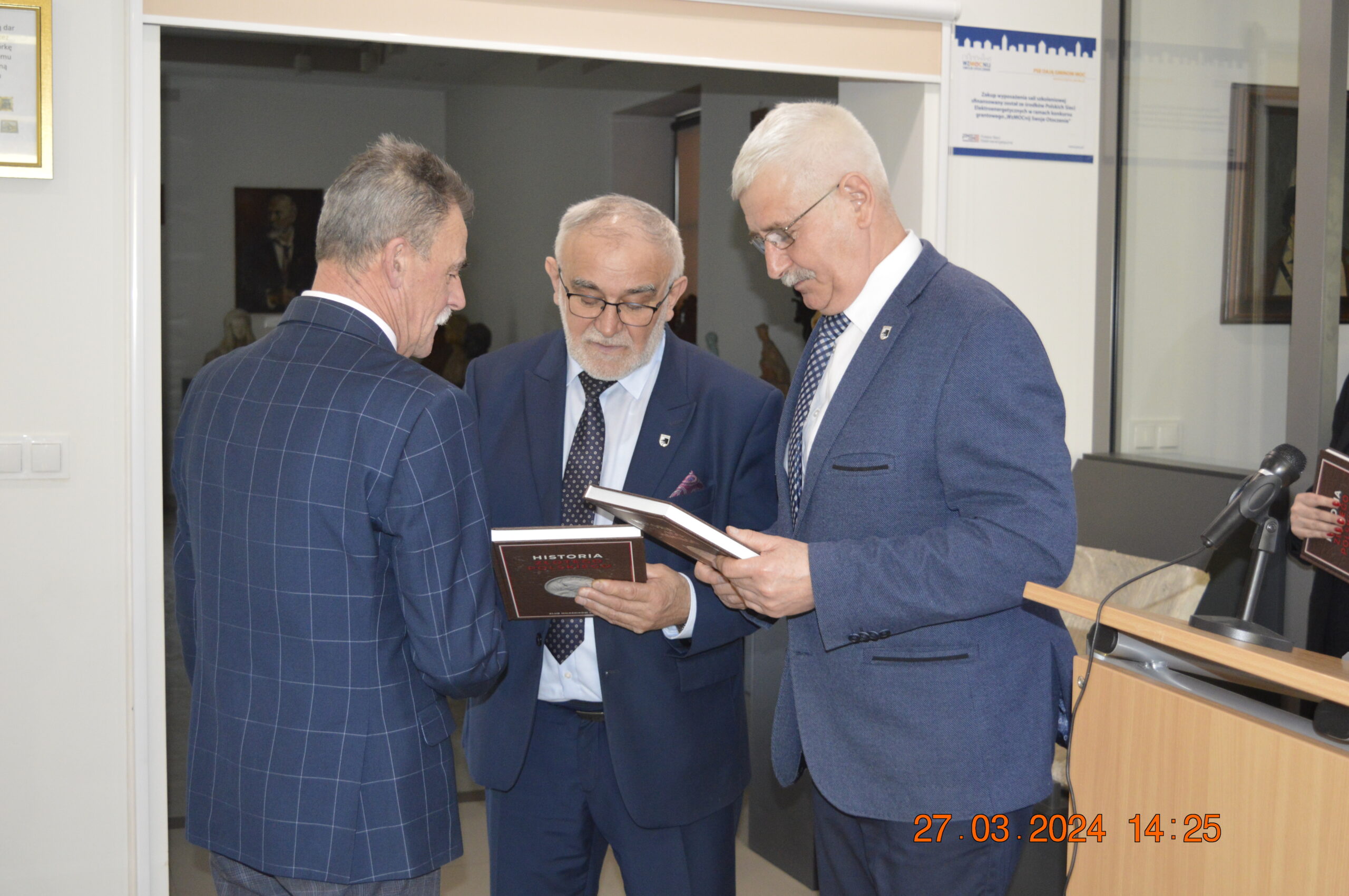 Na obrazku znajdują się trzej mężczyźni ubrani w garniturach, którzy rozmawiają ze sobą, stosując i przeglądając dokumenty. Jeden z mężczyzn stojących przed kamerami, podczas gdy dwa zwrócone są w jego stronie.
