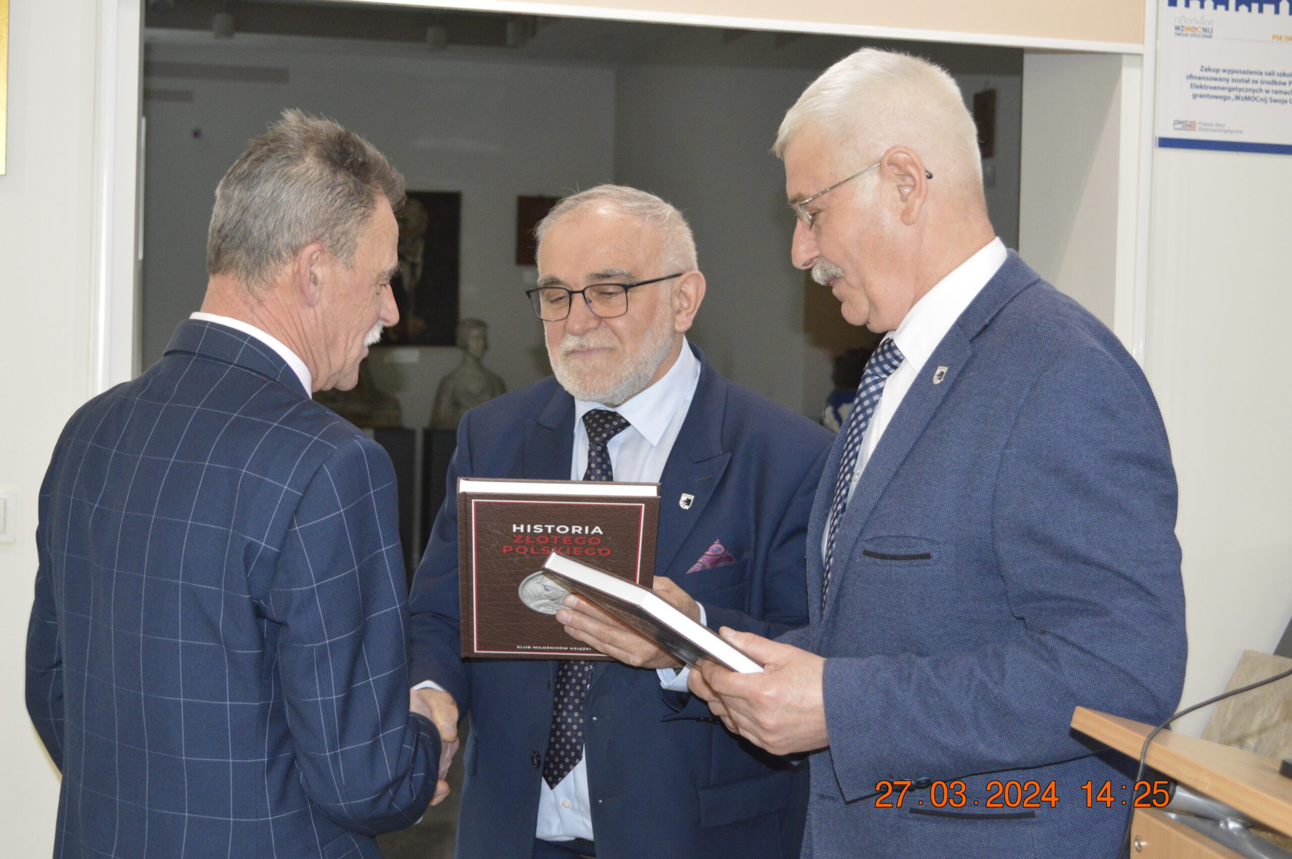 Na obrazku trzech mężczyzn w garniturach wymieniających się książkami wewnątrz pomieszczenia, przy ladzie; jeden z nich, wyciągany na powitanie. W osobnym dolnym rogu widnieje dane i czas "27.03.2024 14:25".
