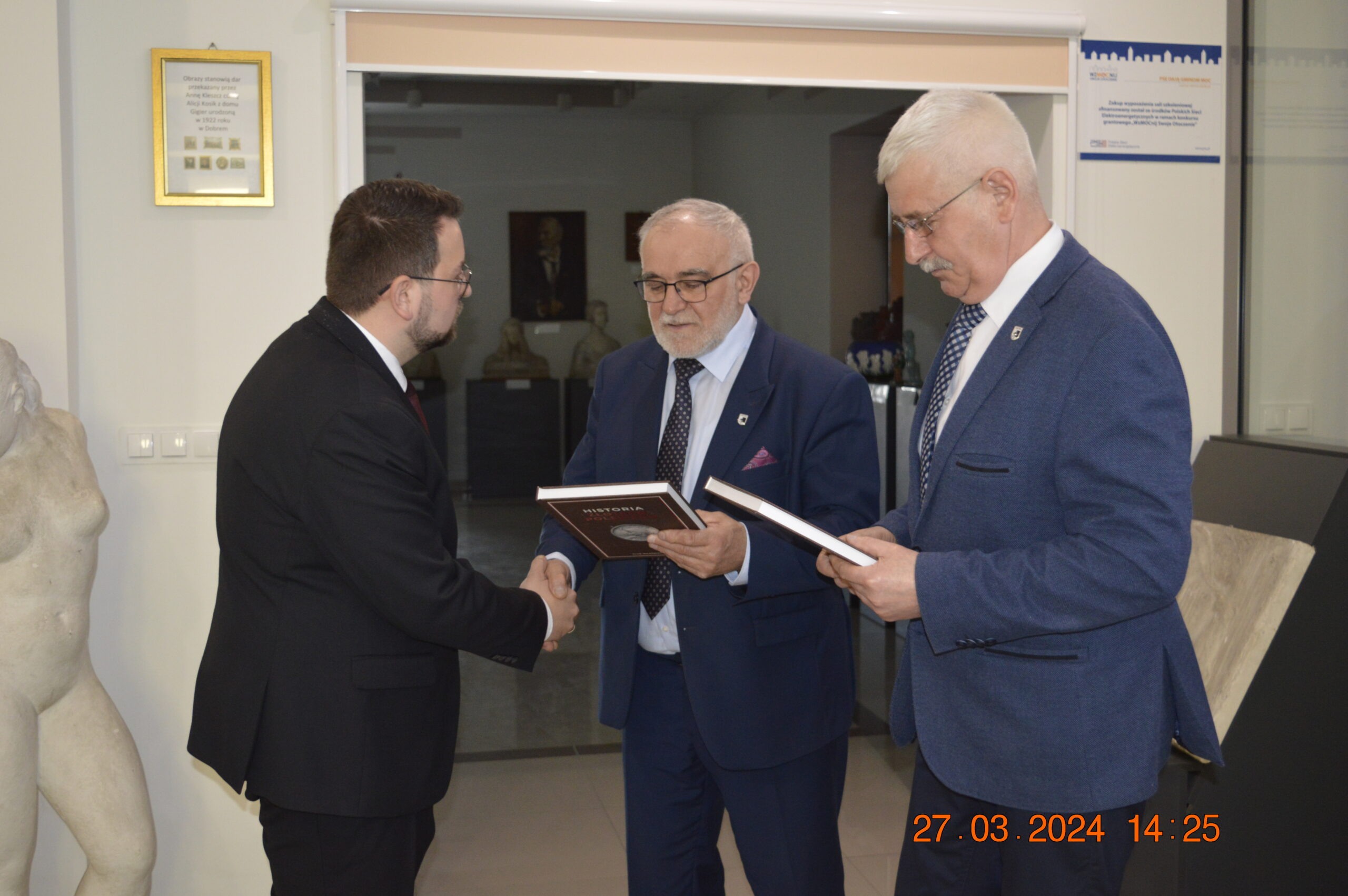 Na obrazku znajdującym się trzech mężczyzn w garniturach w formalnym otoczeniu. Dwóch z nich trzyma twardookładkowe książki, a jeden z nich ściska dłoń trzeciegomu mężczyźnie, przy czym w rogu jest data i godzina „27.03.2024 14:25”.