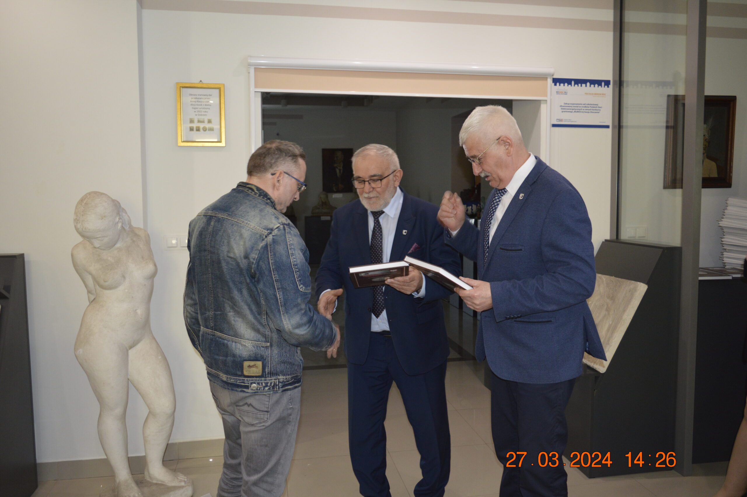 Na obrazku mężczyzny w dżinsowej kurtce podaje jednemu z dwóch mężczyzn w garniturach, którzy trzymają się i certyfikat. Scena rozgrywająca się w dniu, a w tle pojawi się naga rzeźba, przy czym dane do 27.03.2024.