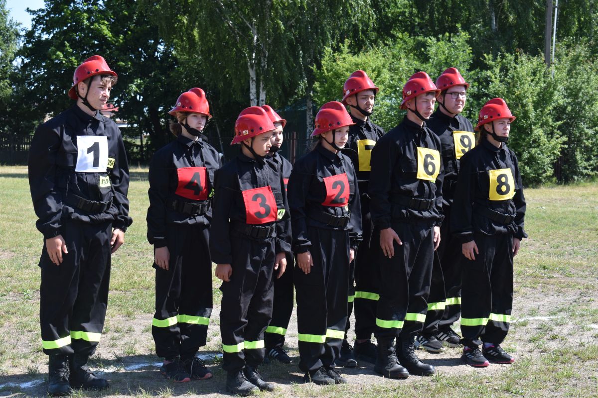 Na obrazie pojawia się grupa osób ubranych w czarne mundury i czerwone hełmy z numerami na piersiach, stojących razem na trawie. W tle widoczne drzewa, co istotne, że są na świeżym powietrzu.