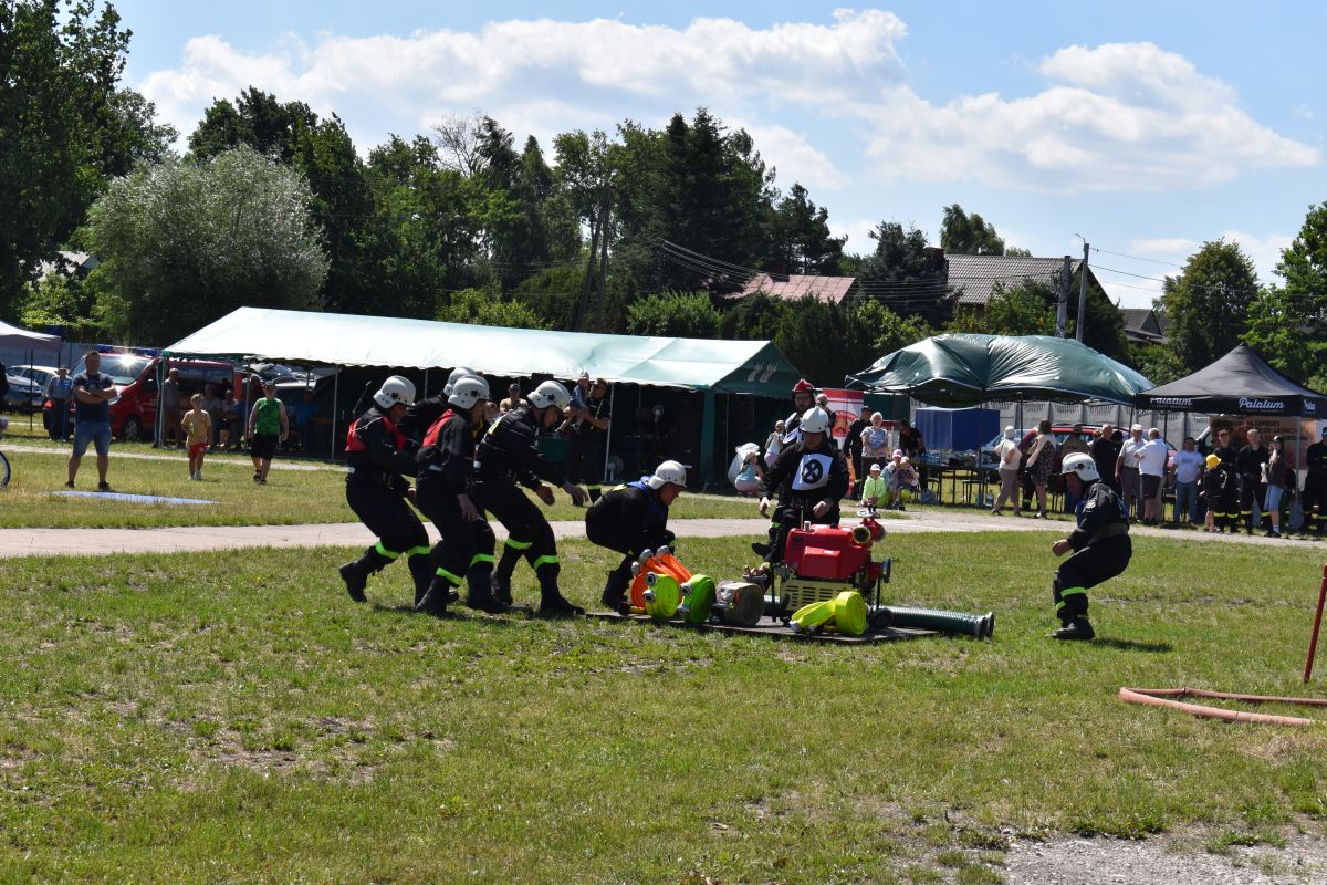 Na zewnątrz znajduje się zespół, który jest w mundurach, który ma udział w działaniach na zewnątrz, obsługując sprzęt na trawiastym terenie. W tle są namioty oraz widoczny się widzowie.