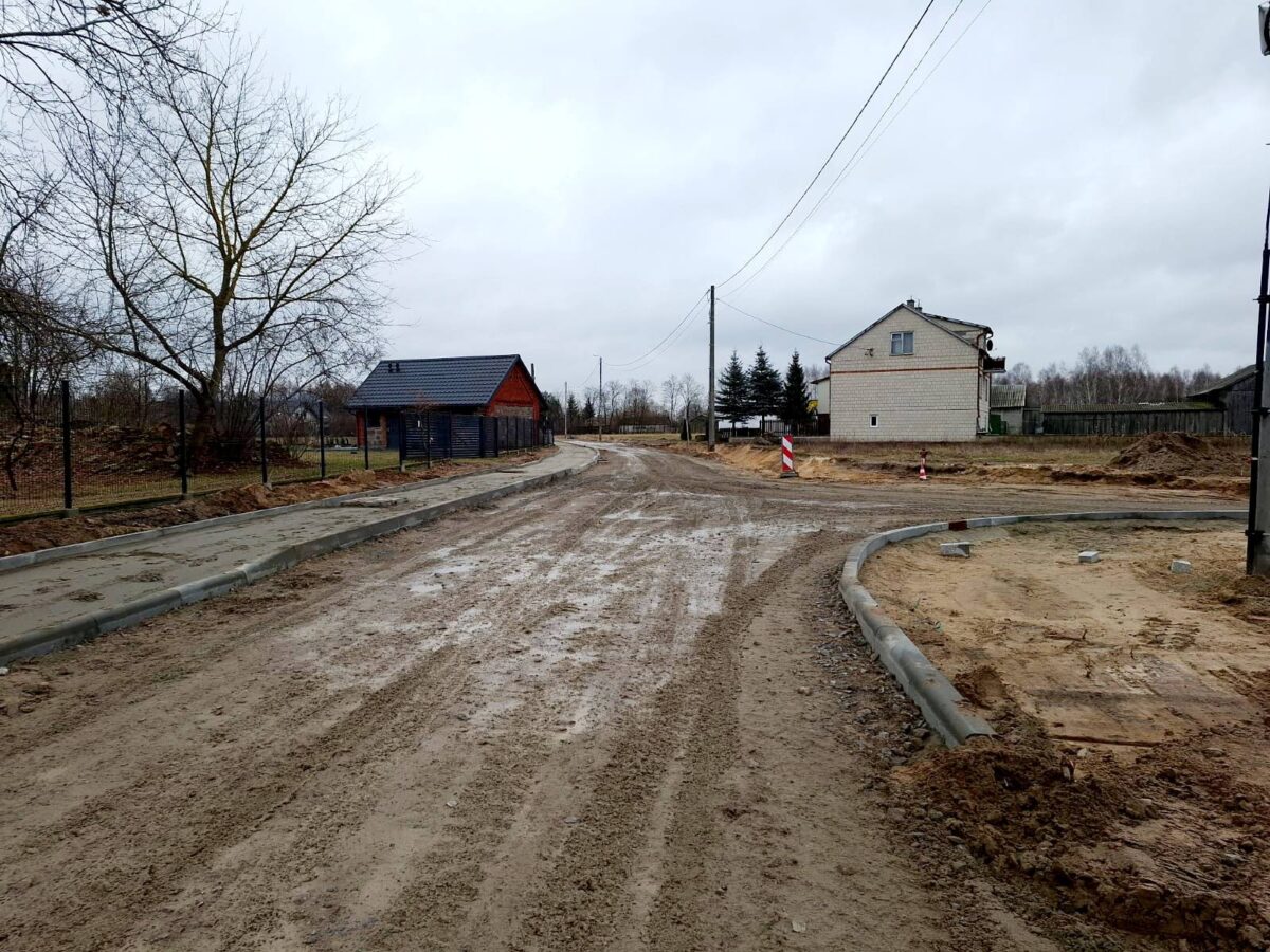 Na obrazku znajduje się błotnista droga w budowie, która może się znajdować na obszarach wiejskich z charakterystycznymi domami i drzewami. utrzymujący się także słupy energetyczne, a całość utrzymana jest w pochmurnej scenerii.