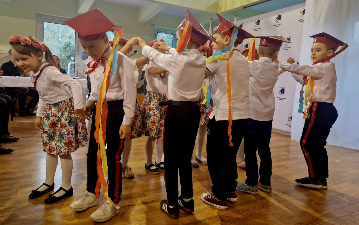 Na obrazku widoczne są dzieci ubrane w kolorowe stroje i czerwone czapki, które słonecznie tańczą trzymając się za ręce oraz trzymając wstążki. wystąpienie zjawiska na drewnianej instalacji w jasnooświetlonym pomieszczeniu.