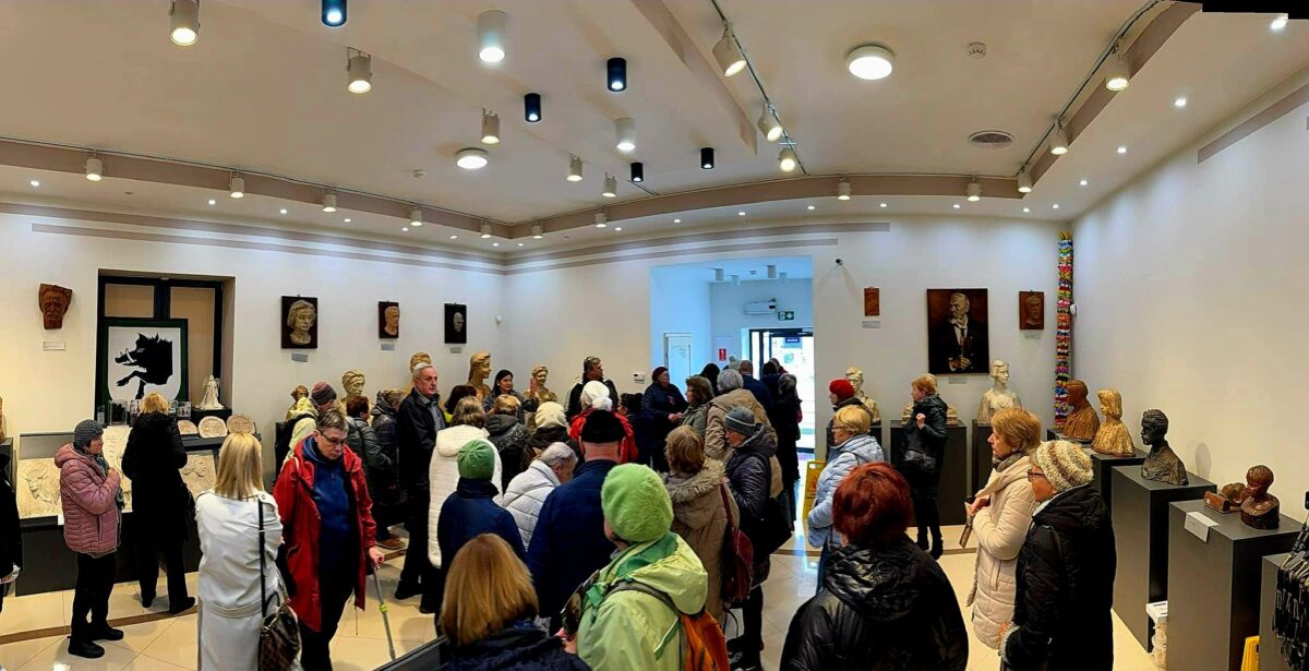 Grupa ludzi w kolorowych kurtkach w sali wystawowej z rzeźbami. Jasne ściany ozdobione popiersiami i obrazami. Nowoczesne oświetlenie sufitowe.