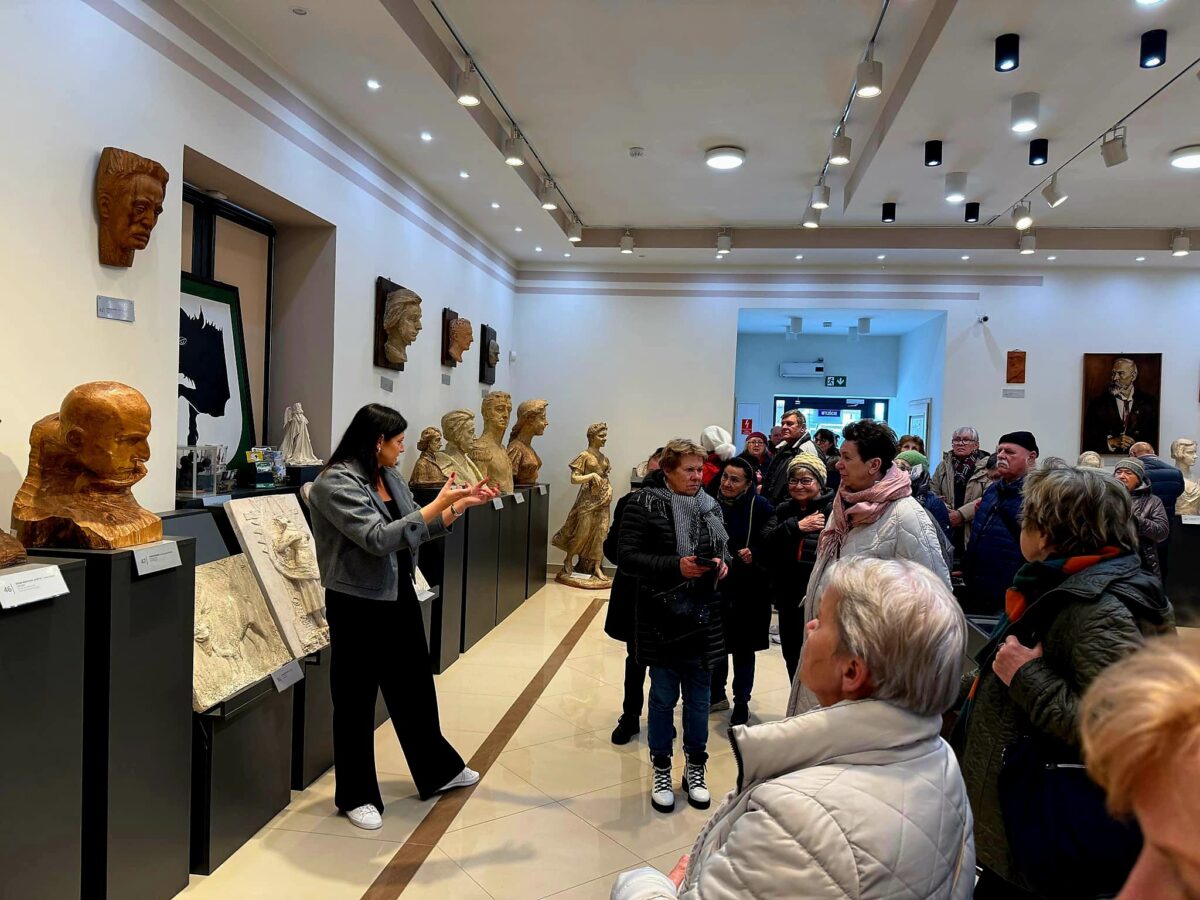 W jasnej galerii prowadzone są zajęcia z rzeźbami na ścianach. Grupa ludzi stoi, słuchając przewodnika. Wnętrze jest eleganckie z intensywnym oświetleniem.