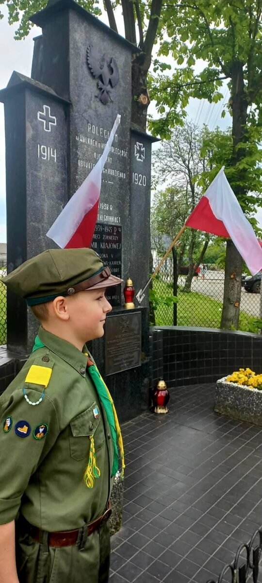 Chłopiec w harcerskim mundurze stoi przed pomnikiem z polskimi flagami. Monument jest ciemny, z białym napisem, tło zielone. Obok znajdują się znicze i kwiaty.