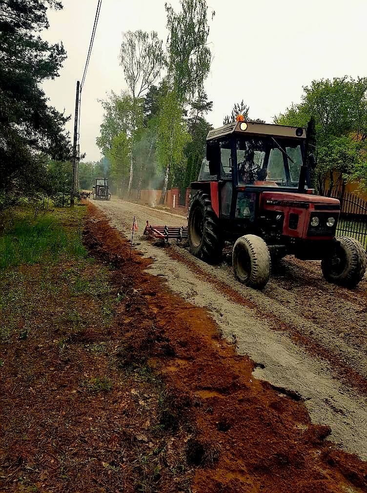 Traktor jedzie po nieutwardzonej drodze w lesie; ciągnie urządzenie do wyrównywania. W tle drzewa i zielone krzewy.