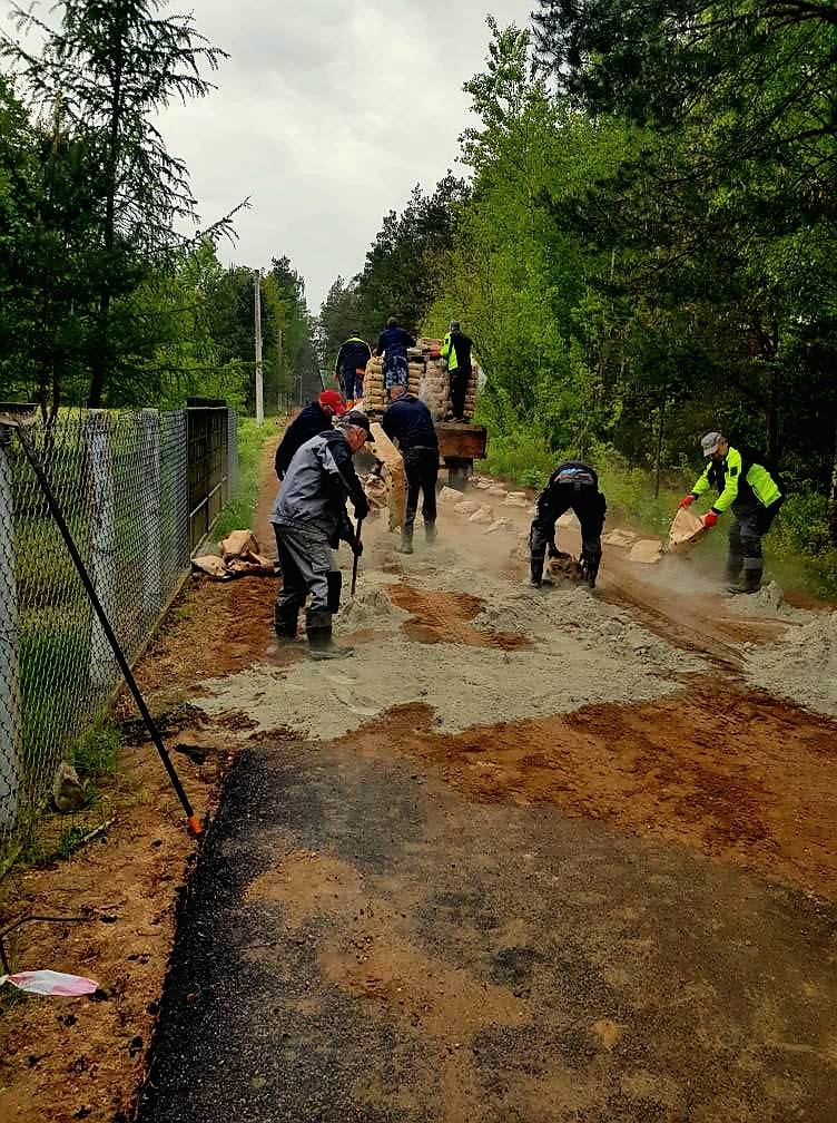 Grupa ludzi pracuje przy naprawie drogi, układając beton. W tle drzewa i chmurne niebo. Po lewej ogrodzenie.