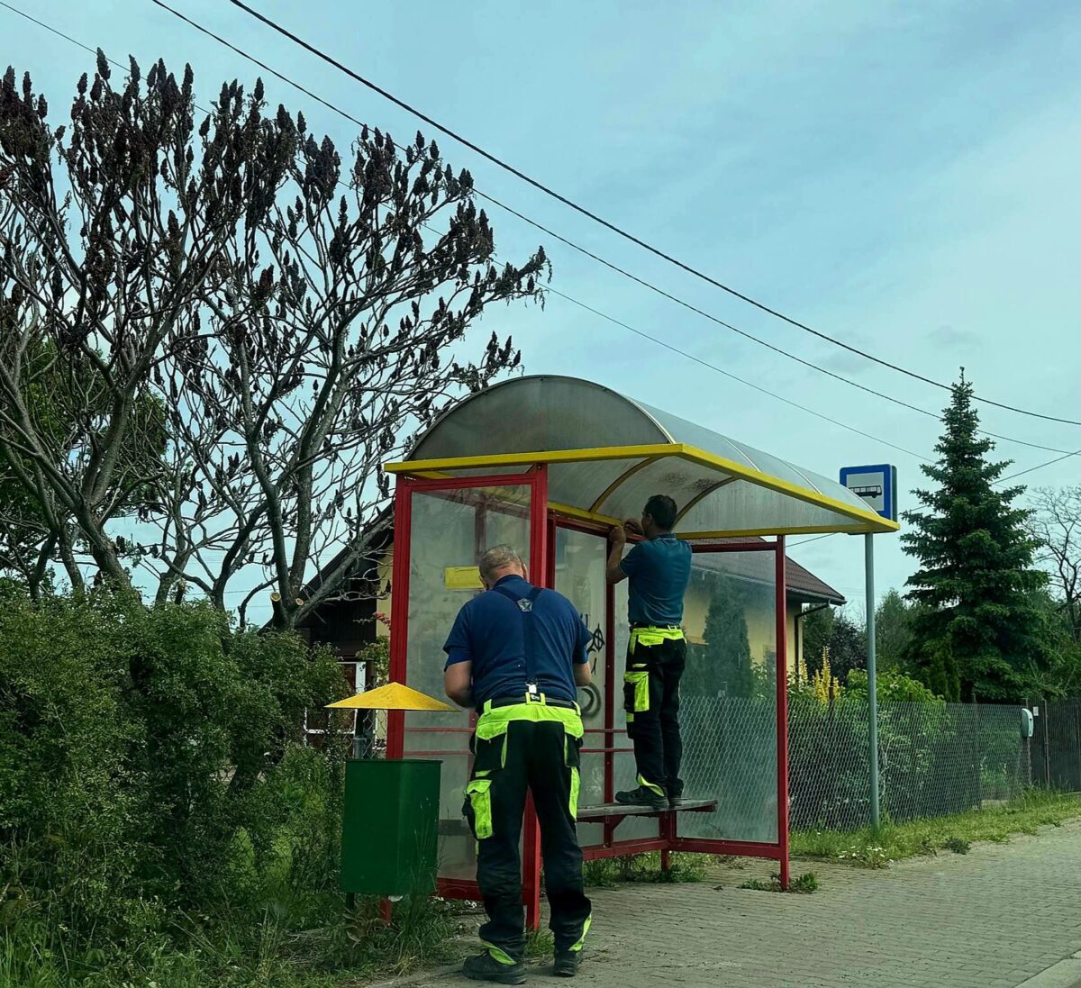 Dwóch pracowników w odblaskowych strojach naprawia przystanek autobusowy z żółtym dachem na tle zieleni i nieba.