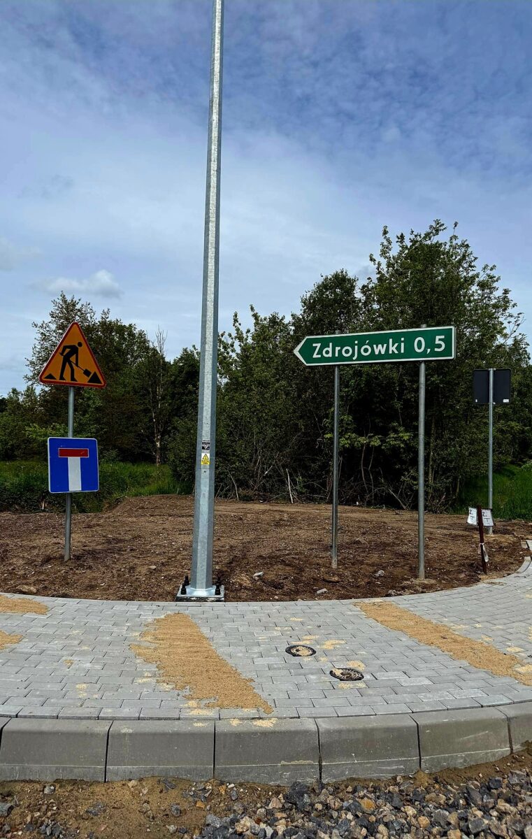 Znak drogowy wskazuje drogę do Zdrojówek, obok znak robót. Na tle drzewa, chodnik i lampa. Dzień jest pochmurny.