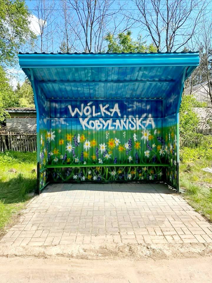 Niebieska wiata przystankowa z napisem Wólka Kobylańska. Tło ozdobione kolorowymi kwiatami. Otoczona zielenią i drewnianym płotem.