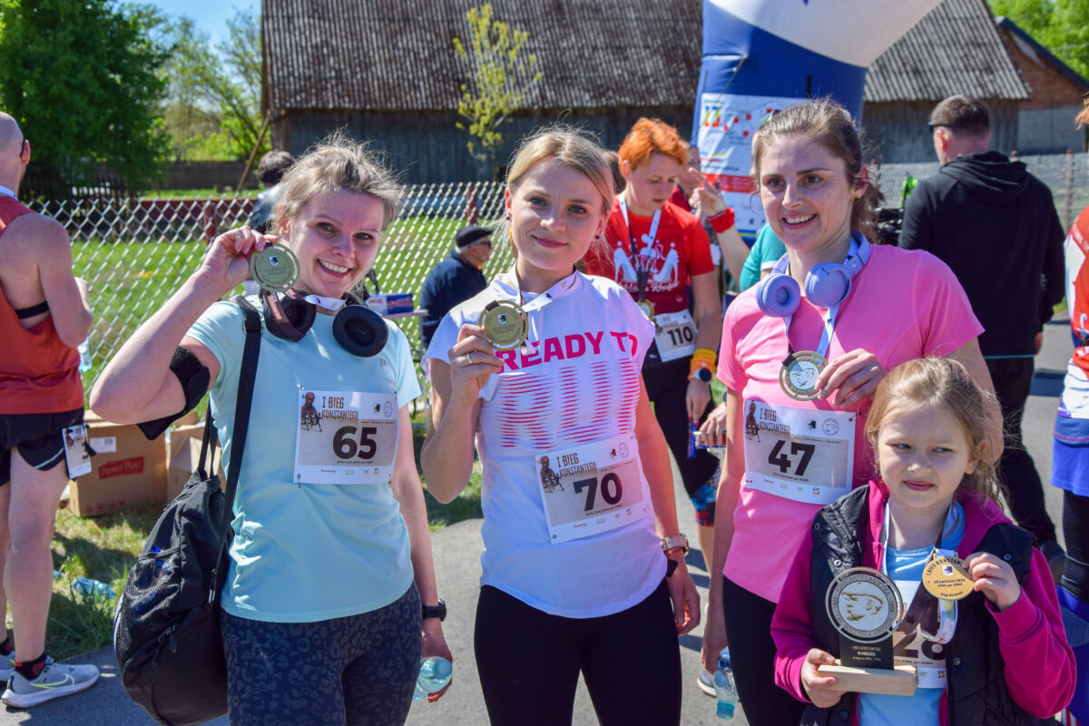 Trzy kobiety z medalami po biegu, w tle zielone drzewa i budynek. Mają stroje sportowe w jasnych kolorach. Uśmiechnięte, podniosły puchary.