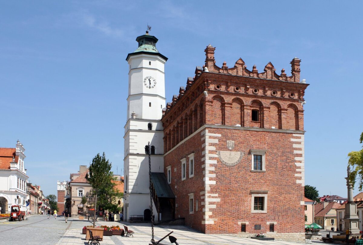 Ceglany budynek z białą wieżą zegarową na tle błękitnego nieba. Otoczony brukowanym placem z drzewami. Architektura renesansowa.