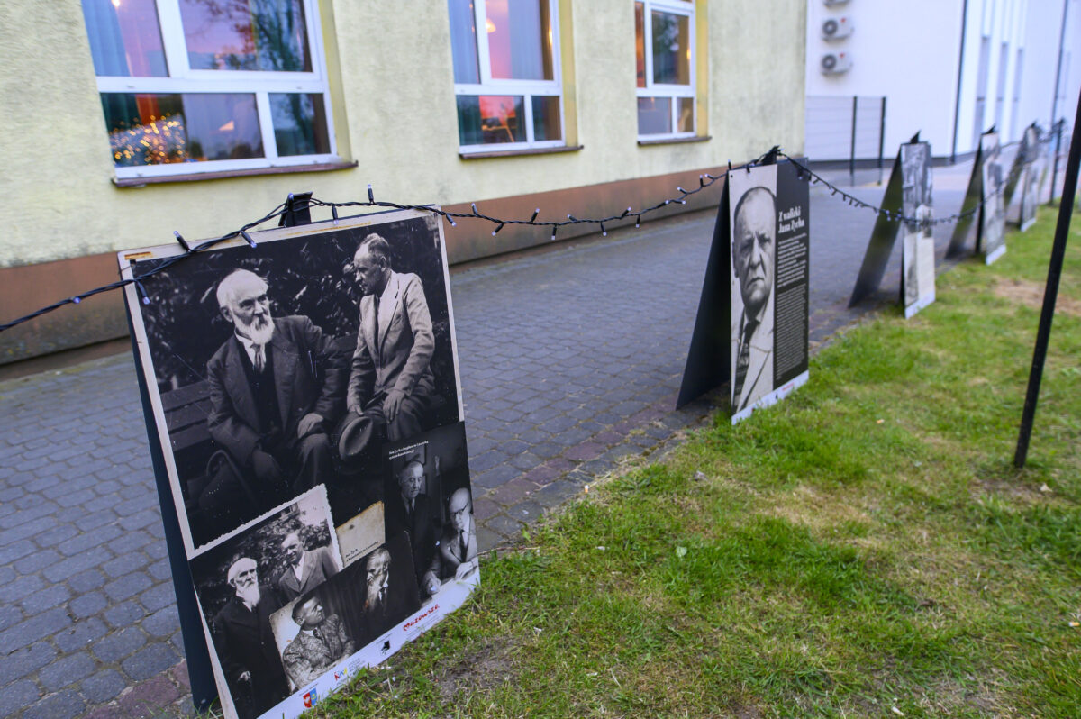 Na zdjęciu widać wystawę plenerową z czarno-białymi fotografiami na tle ściany budynku. Fotografie są ustawione wzdłuż chodnika, ozdobione światełkami. Na trawie obok ustawione są tablice z opisami.