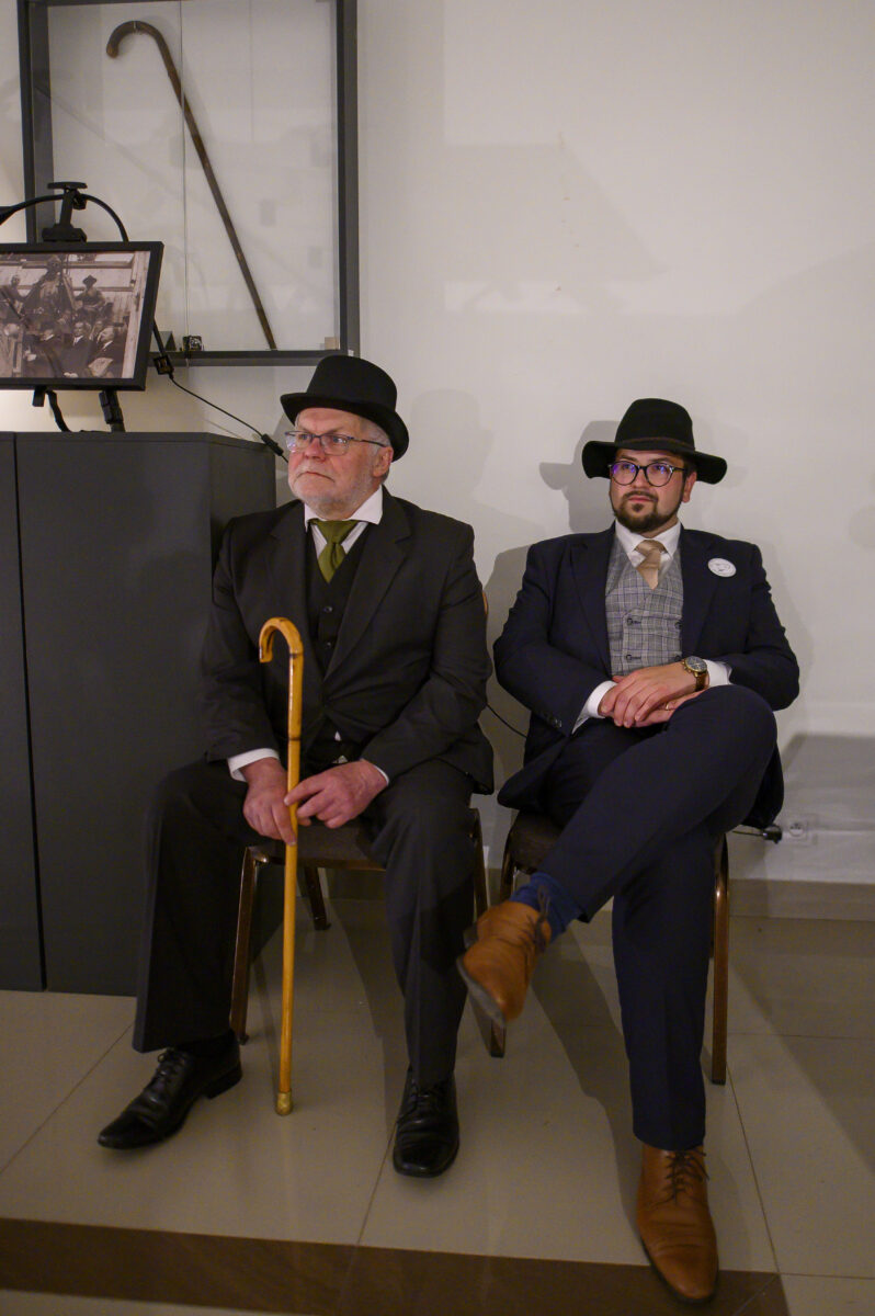Dwóch mężczyzn w eleganckich garniturach i kapeluszach siedzi na krzesłach; jeden trzyma żółtą laskę. Tło białe, obok fotografia i laska w gablocie. Mają skupiony wyraz twarzy.