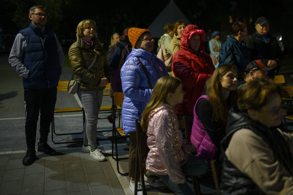 Grupa ludzi w zimowych kurtkach stoi na zewnątrz. Kobiety i mężczyźni w kolorowych ubraniach, na tle ciemnego nieba. Sceneria nocna, obok chodnik.