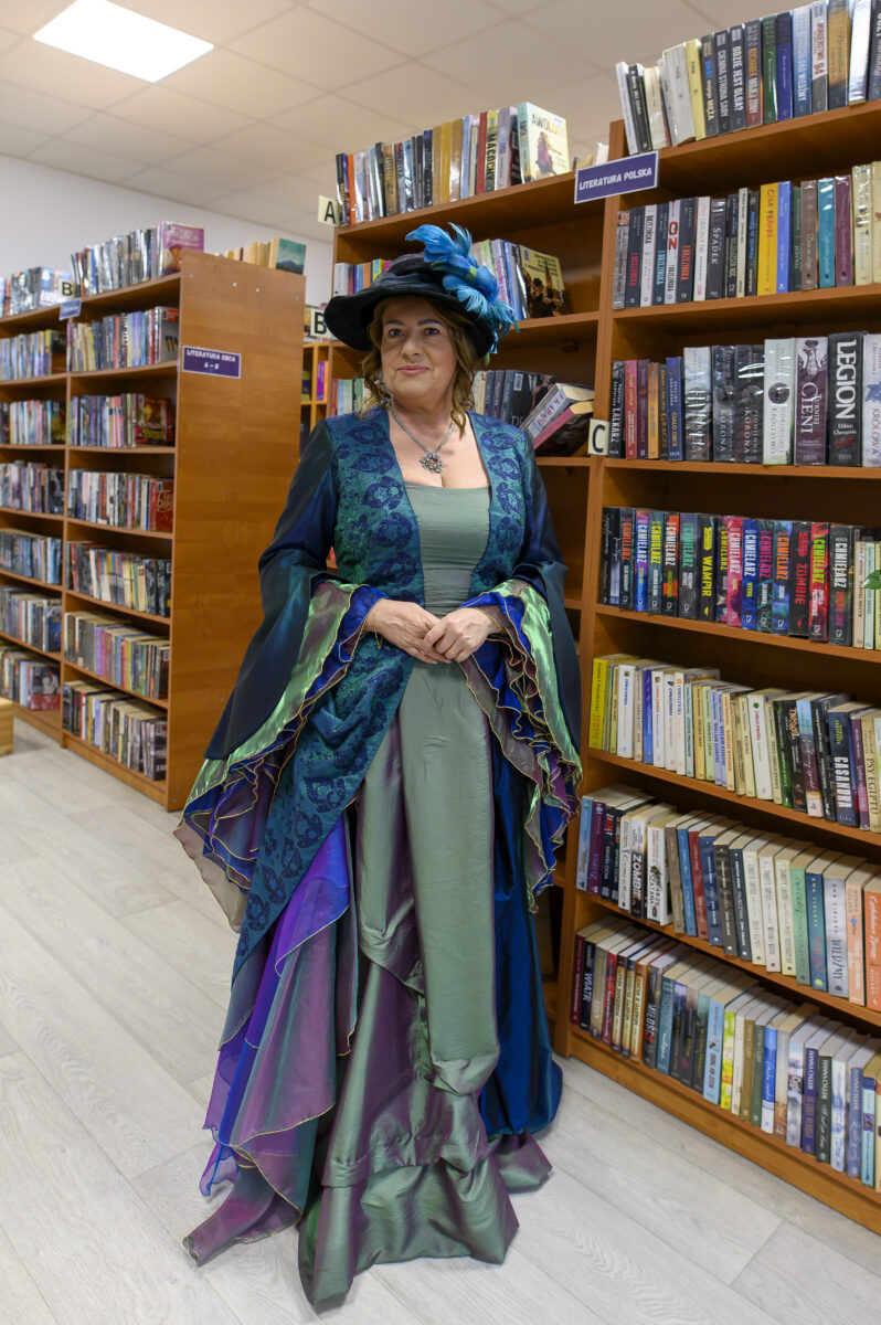 Osoba w eleganckiej sukni z kapeluszem znajduje się w bibliotece. Suknia w zieleni i fioletach harmonizuje z książkami na półkach. Tło stanowią regały z książkami oznaczonymi kategoriami.