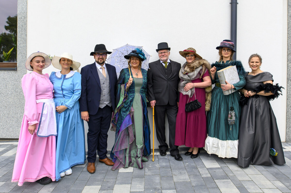 Grupa ludzi w strojach retro pozuje na tle białej ściany. Kolory dominujące to różowy, niebieski i zielony. Kilka osób ma kapelusze.
