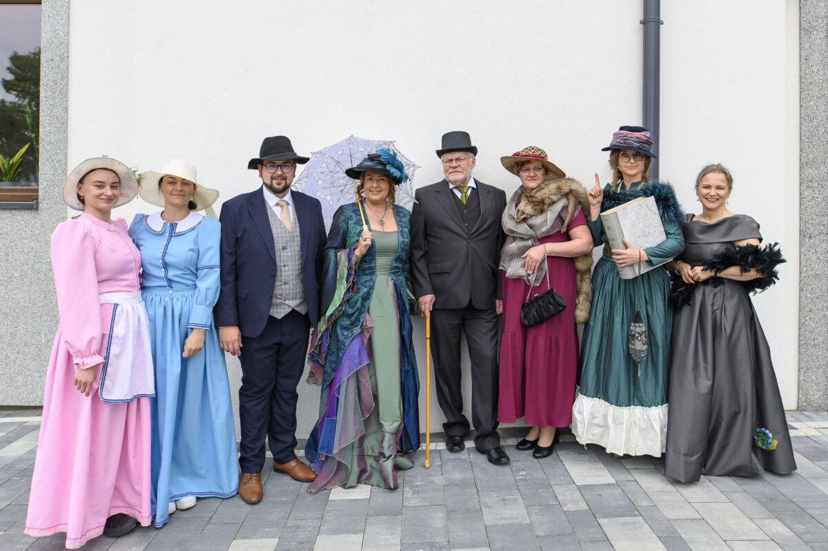 Grupa ludzi w strojach retro pozująca na tle białej ściany. Stroje w kolorach różowym, niebieskim i zielonym. Dwie osoby trzymają parasolkę i książkę.