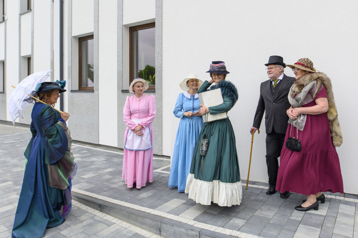 Grupa osób w strojach retro pozuje na tle białej ściany. Kolorowe suknie i kapelusze przyciągają wzrok. Kafelkowana, szara posadzka.