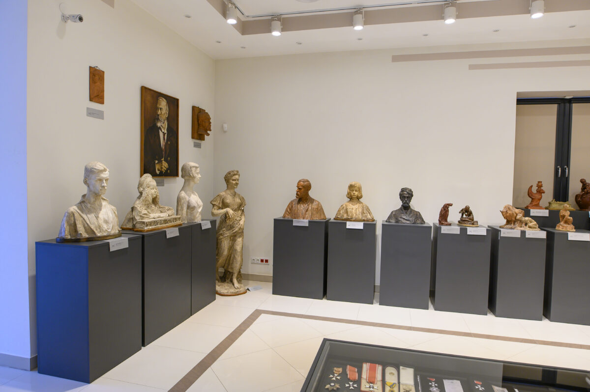 Na zdjęciu widzimy galerię popiersi i rzeźb w jasnym pomieszczeniu. Popiersia stoją na szarych postumentach, a tło jest kremowe. Nad popiersiami wisi obraz portretowy.