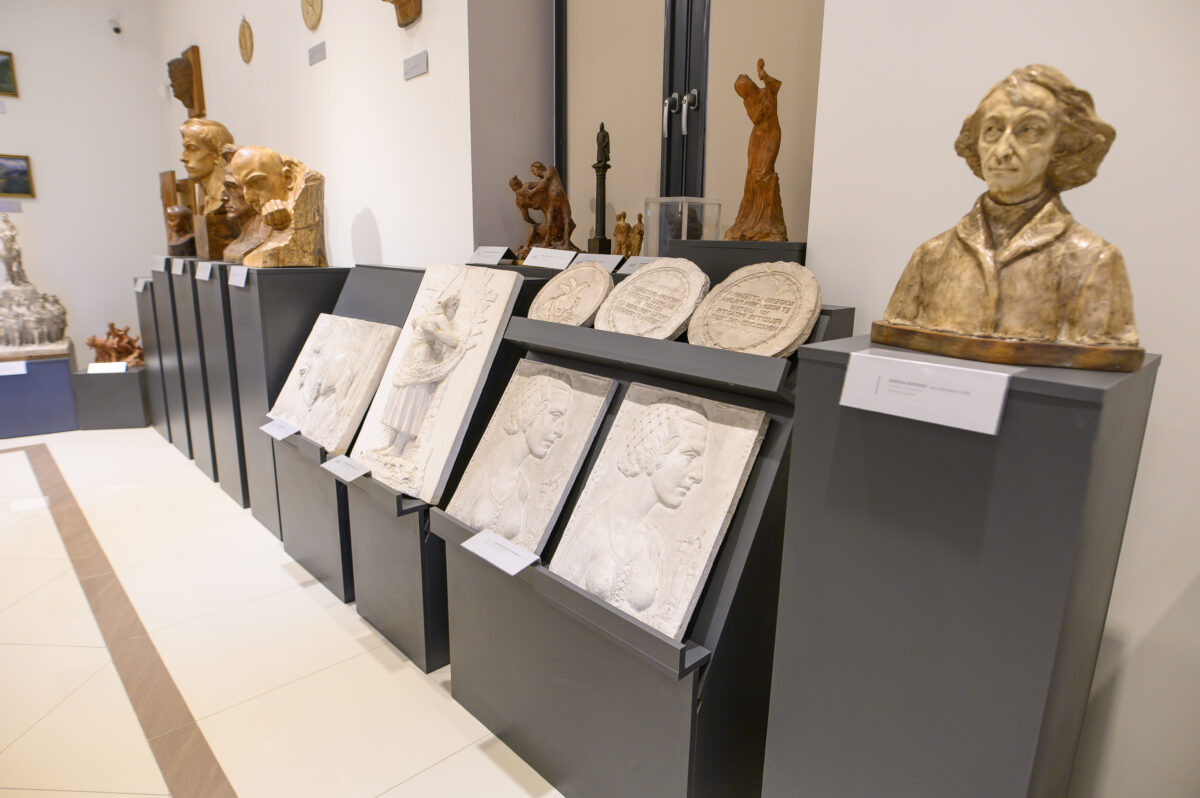Galeria z popiersiami, płaskorzeźbami i rzeźbami na czarnych postumentach. Jasne ściany, kontrastujące ciemne podstawy. Rzeźby w ciepłych odcieniach brązu i bieli.
